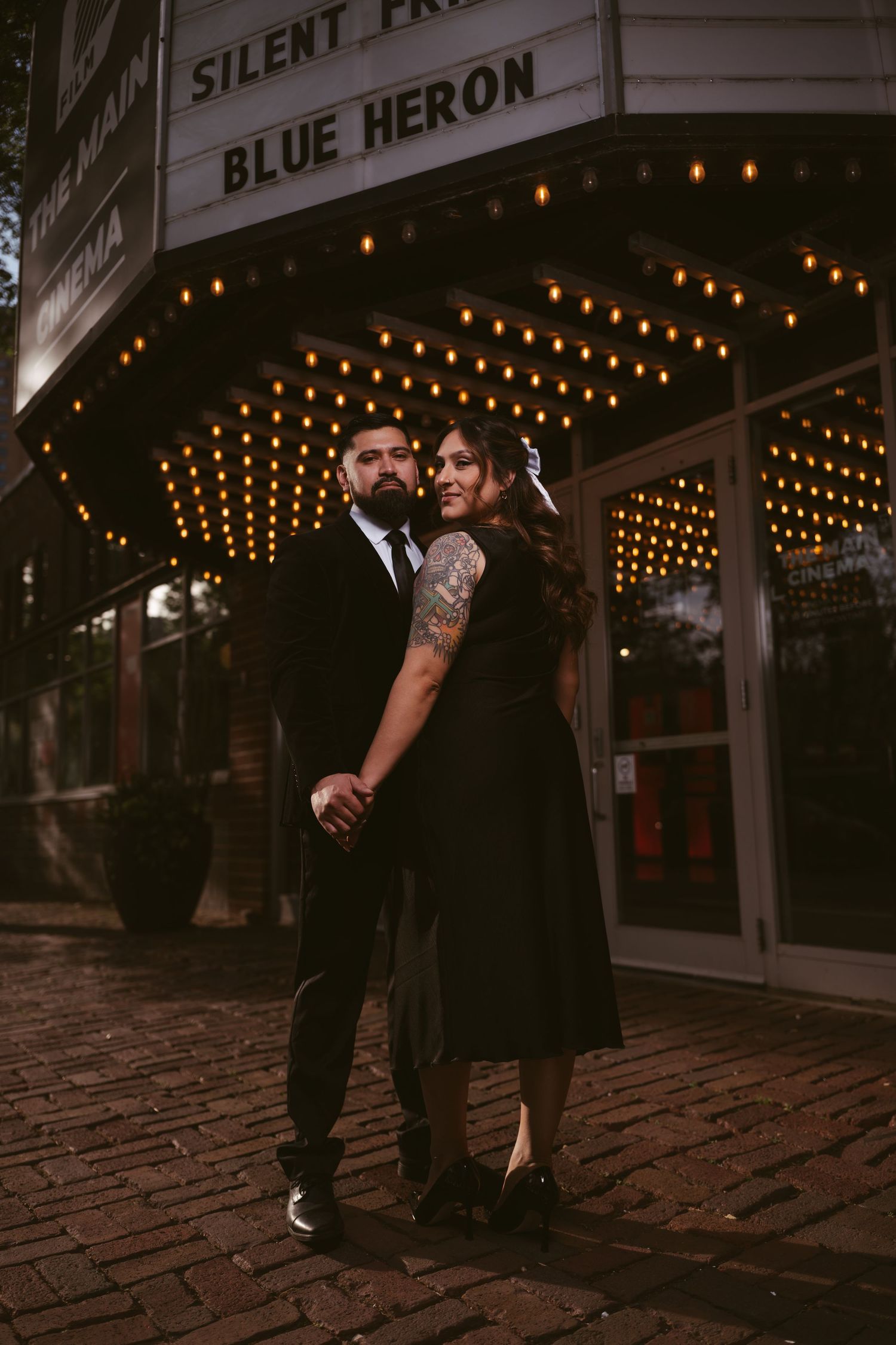 Couple in black formal attire poses hand-in-hand before a lit marquee sign reading Blue Heron at night.