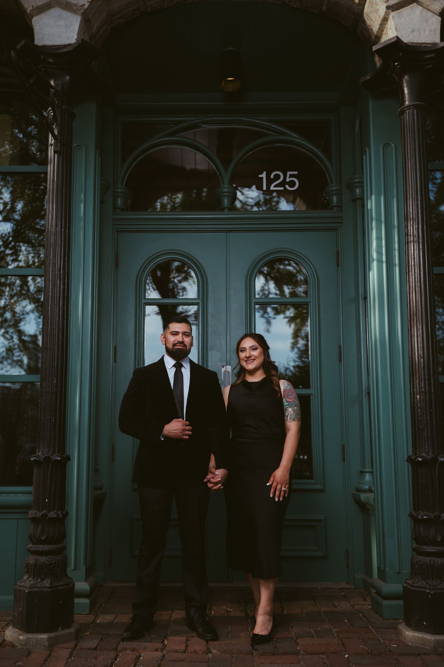 Couple in black attire posing before teal door numbered 125 in urban setting.