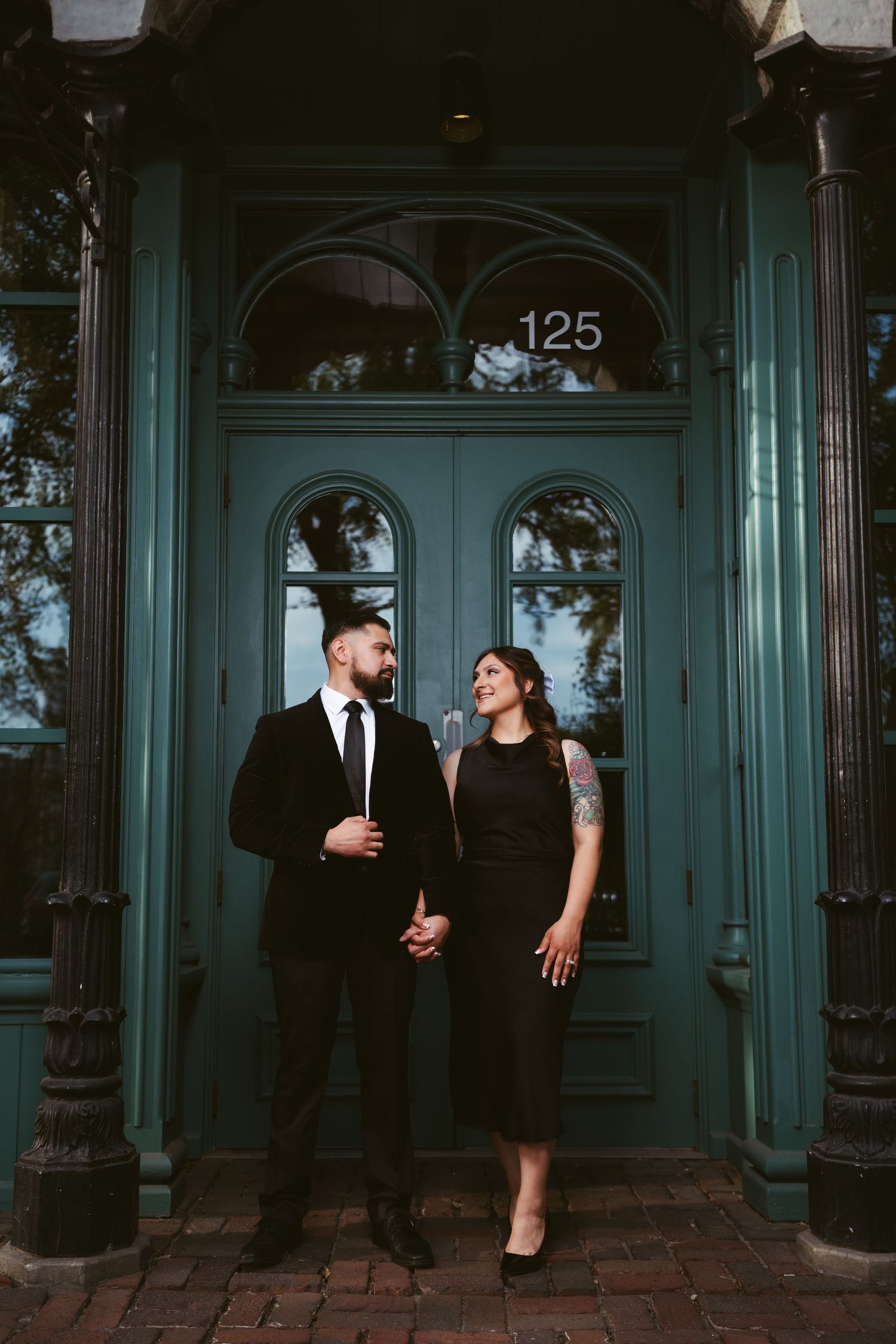 Elegant couple dressed in black stand before a teal door numbered 125, sharing a loving glance.