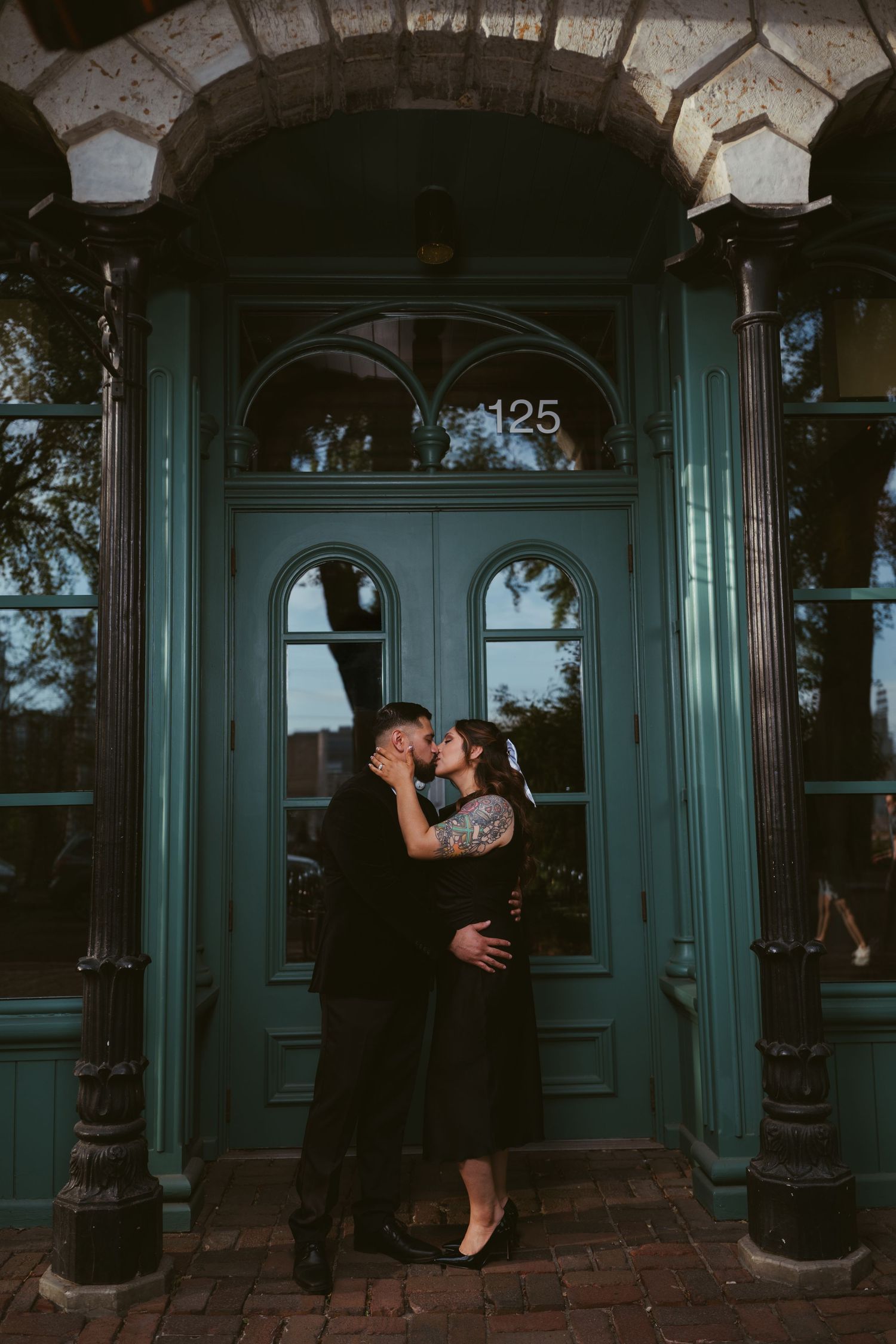 A couple shares a romantic kiss in front of teal double doors numbered 125 at dusk.
