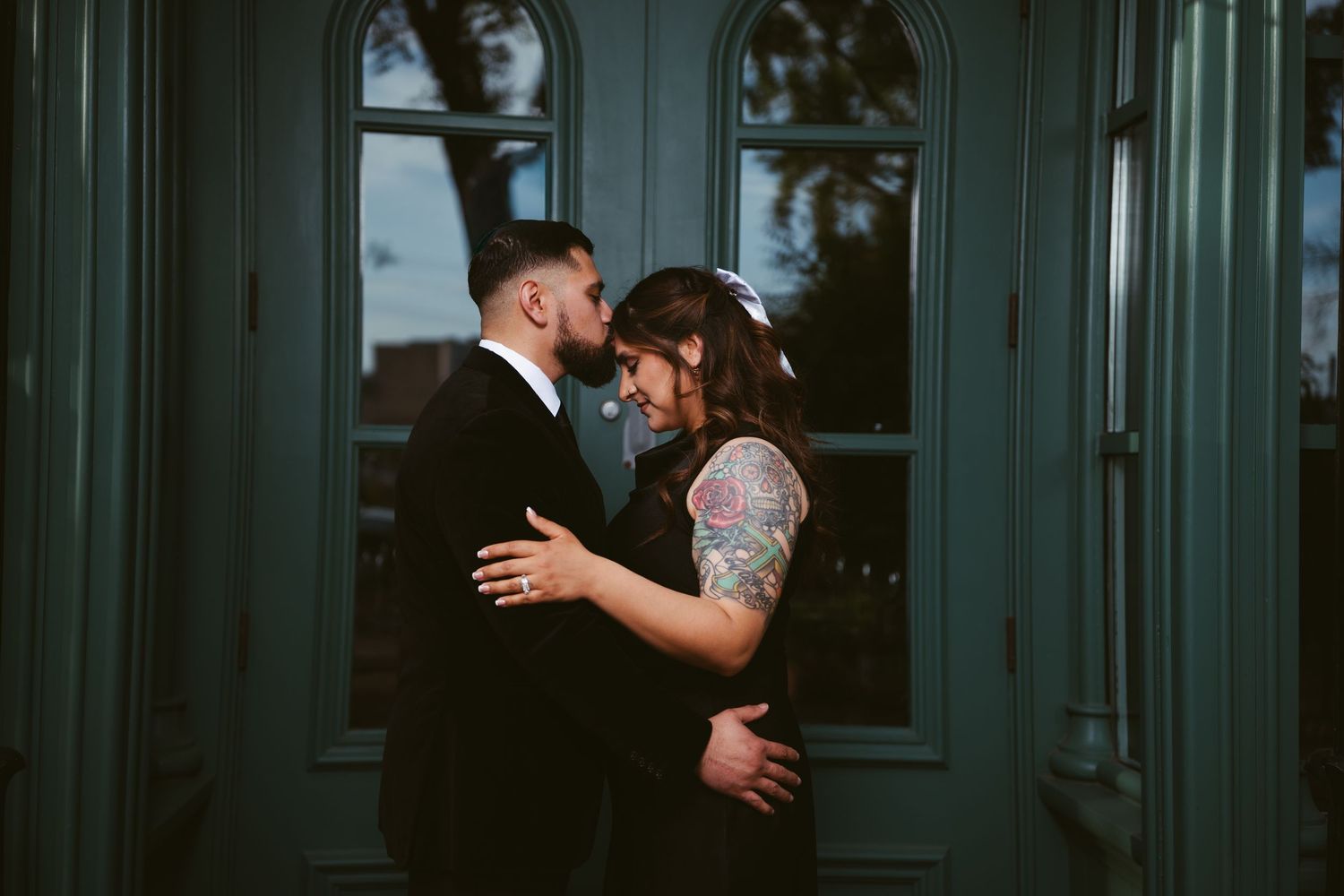 A couple shares a romantic moment in wedding attire, the bride with tattoos and groom in a suit, posed before teal doors.