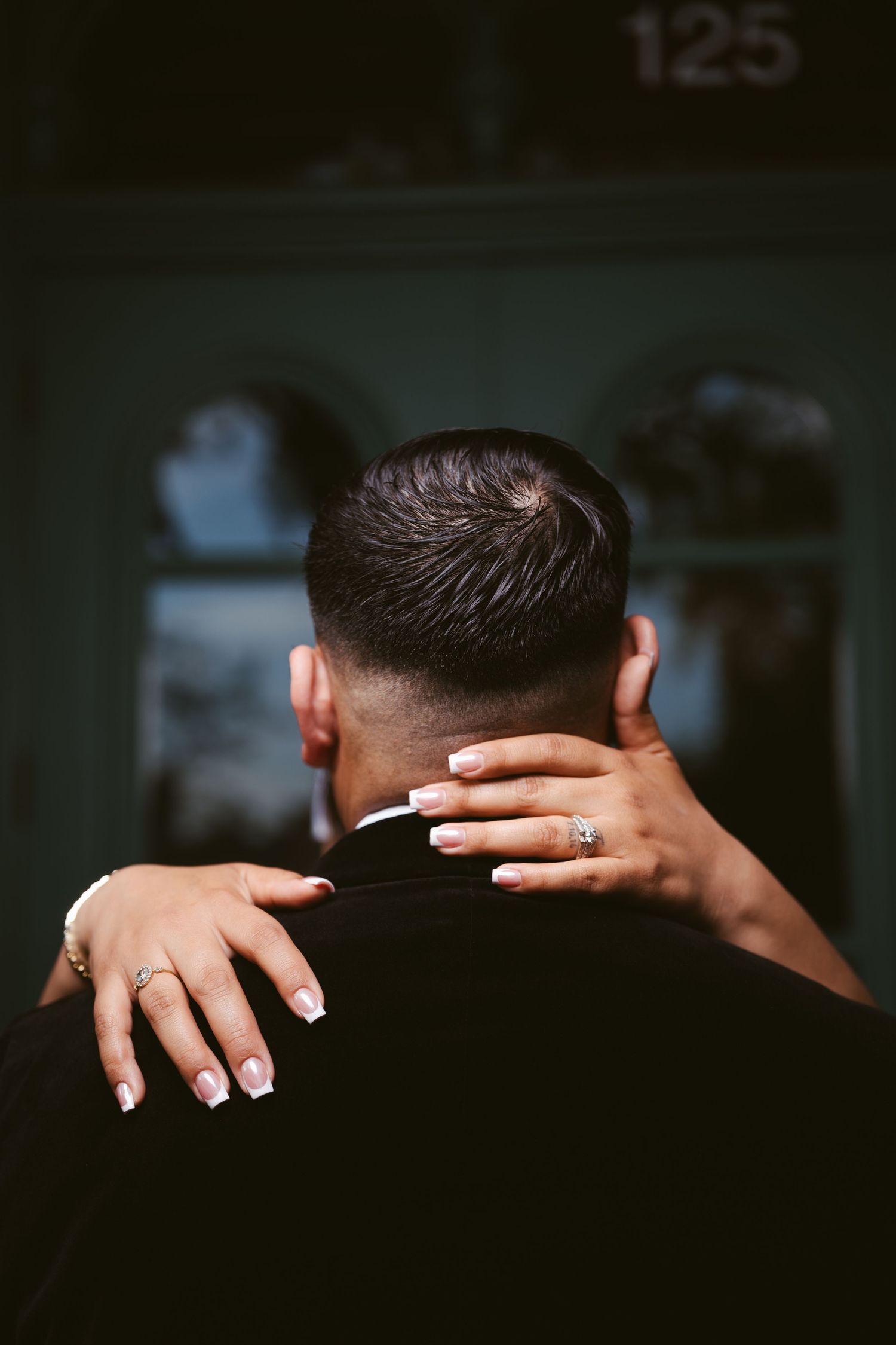 A woman with French manicured nails and a diamond ring embraces a man from behind near a door marked 125.