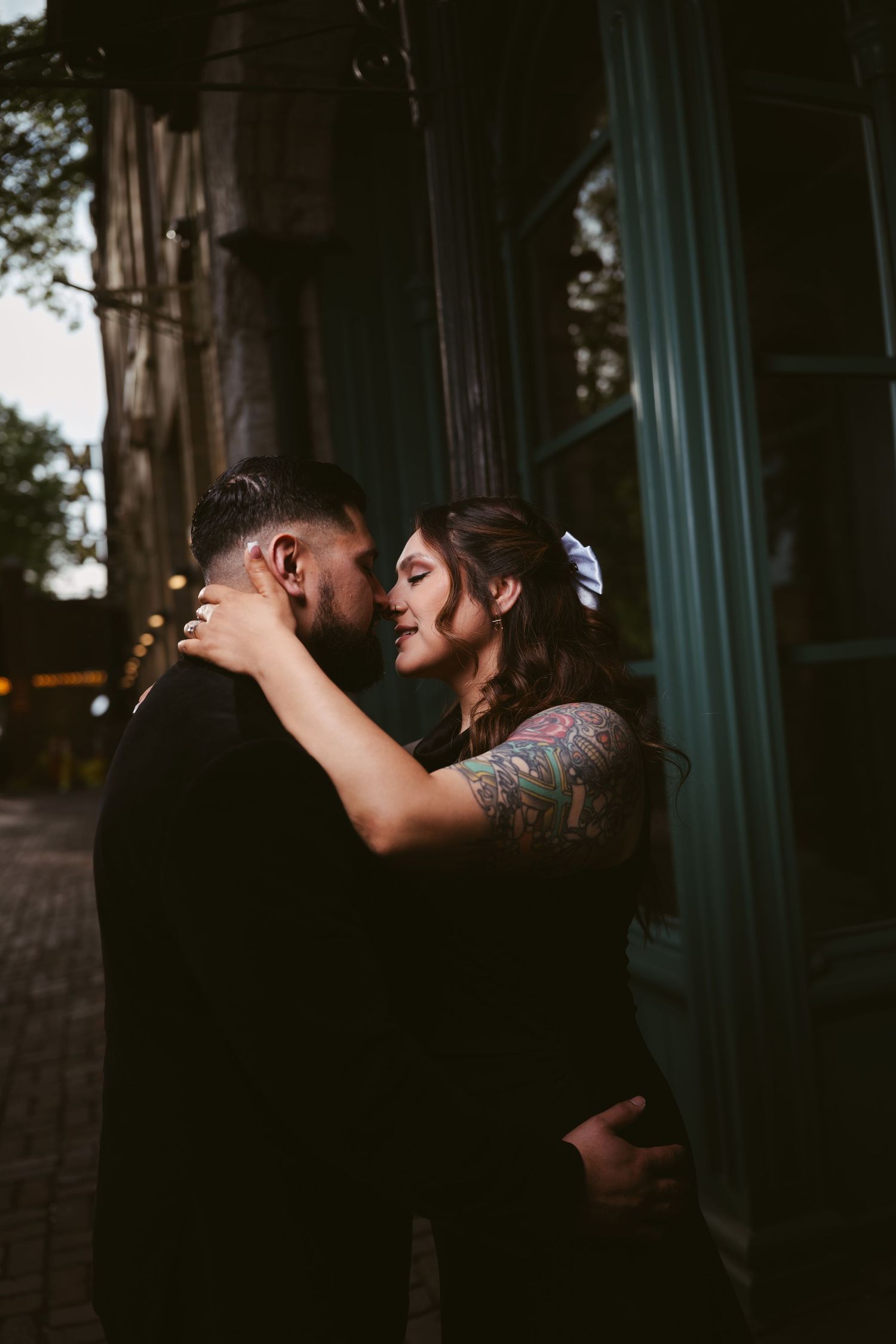 A couple shares a romantic near-kiss embrace outdoors at dusk, the woman adorned with tattoos and a white hair bow.