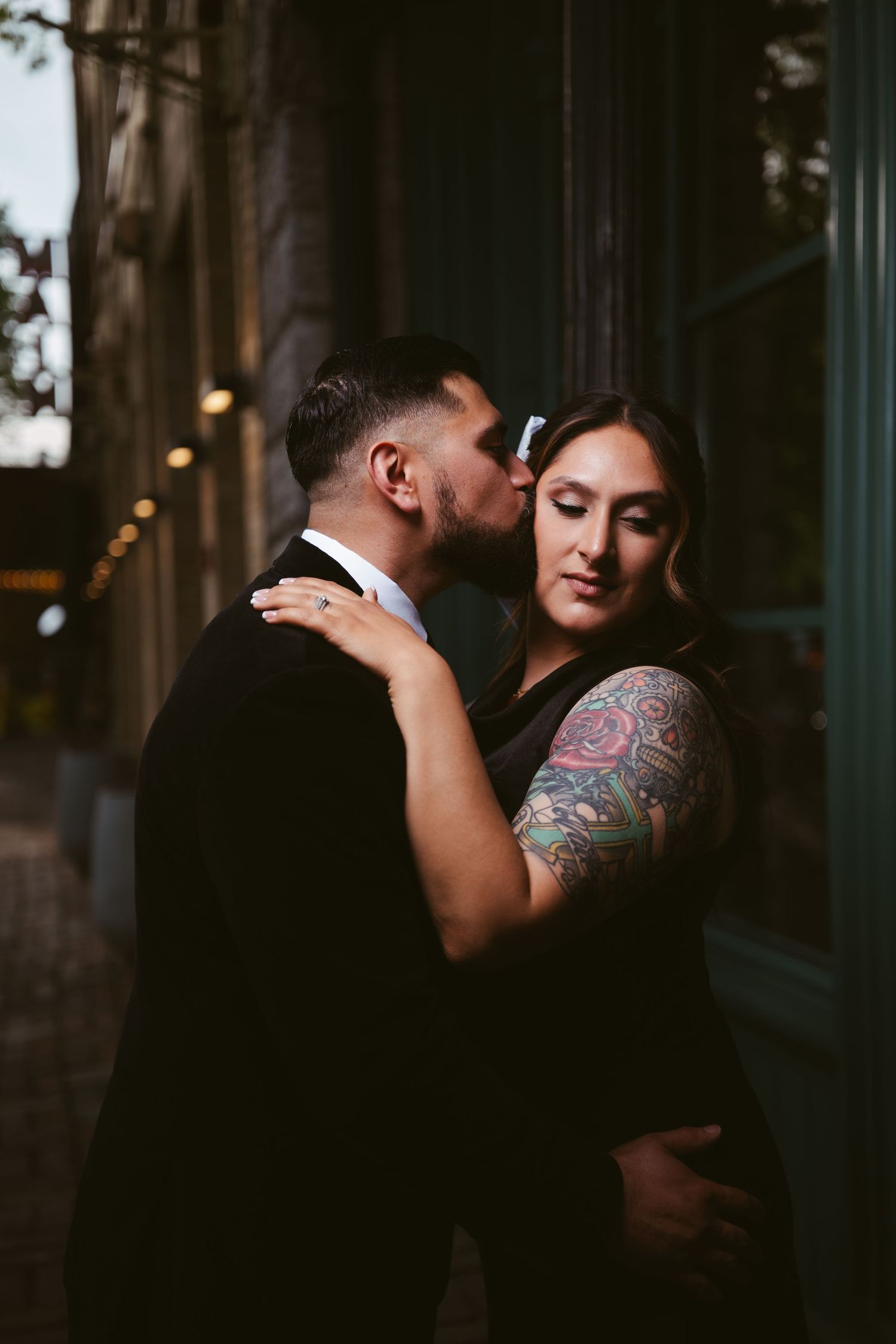 A couple shares a tender moment, man kissing woman with colorful tattoo sleeve, both dressed in black at dusk.