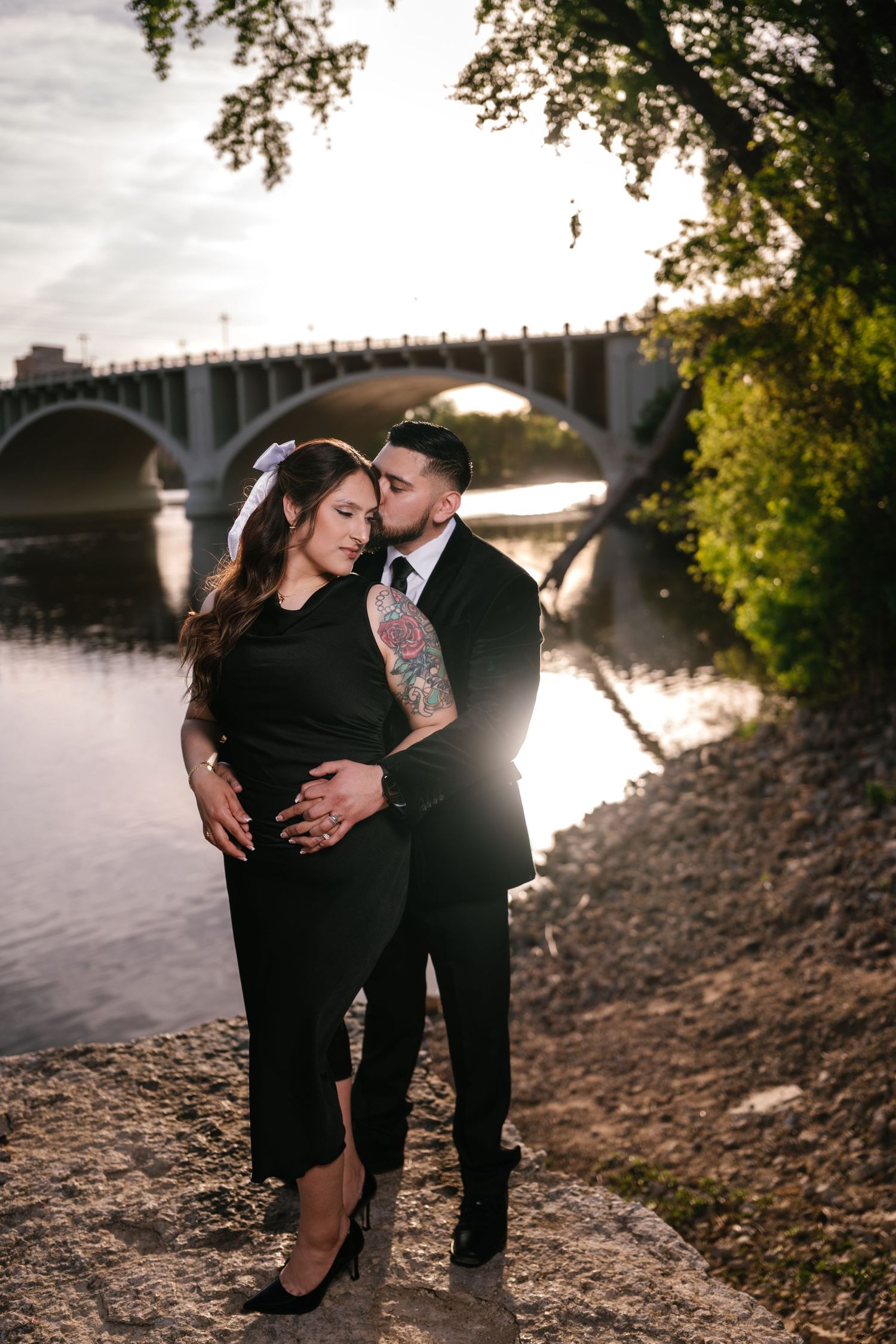 Couple in black formal attire embrace by a sunlit river with a stone arch bridge in the background.
