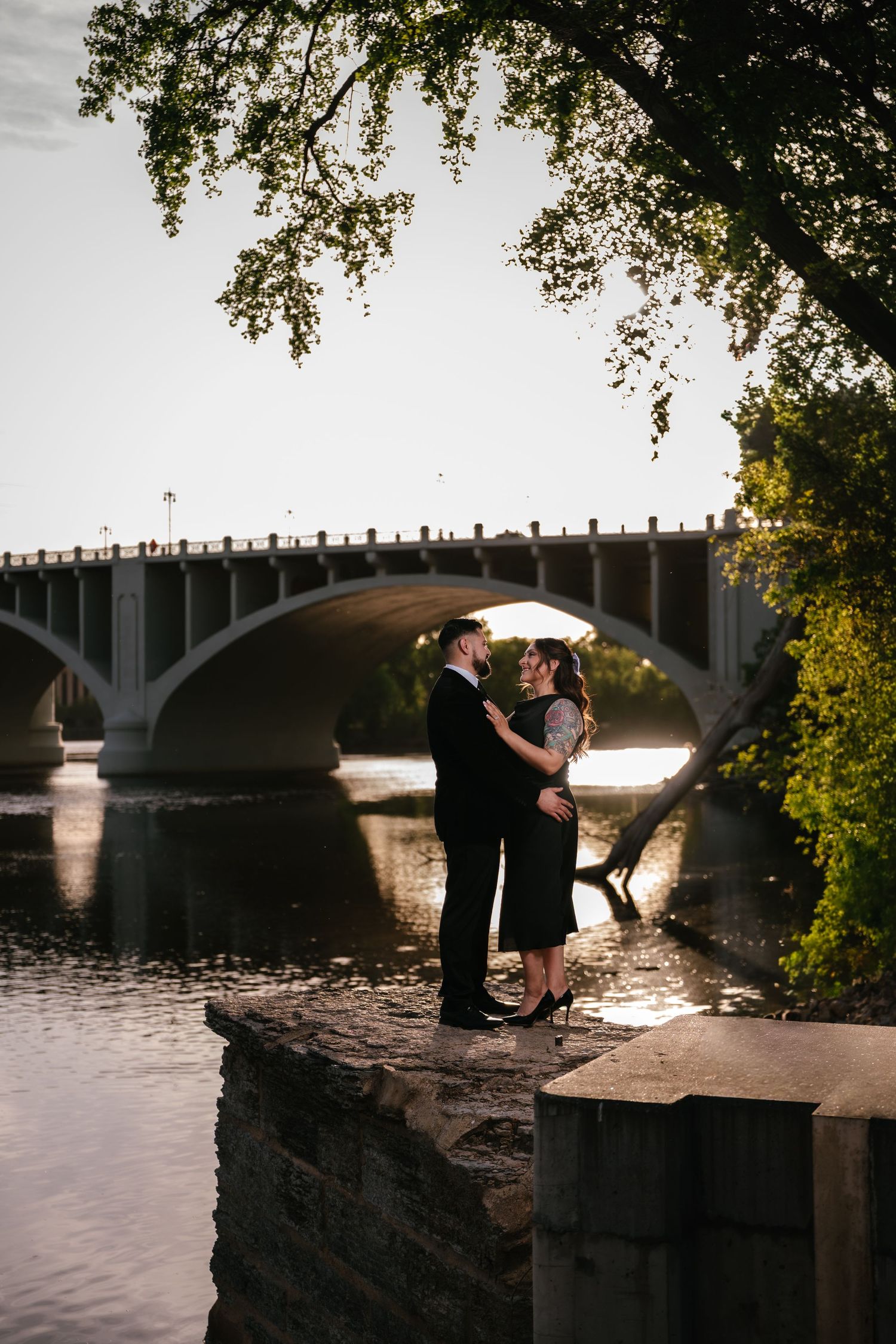 Couple embracing romantically on stone wall by river with arched bridge and golden sunset light behind them.