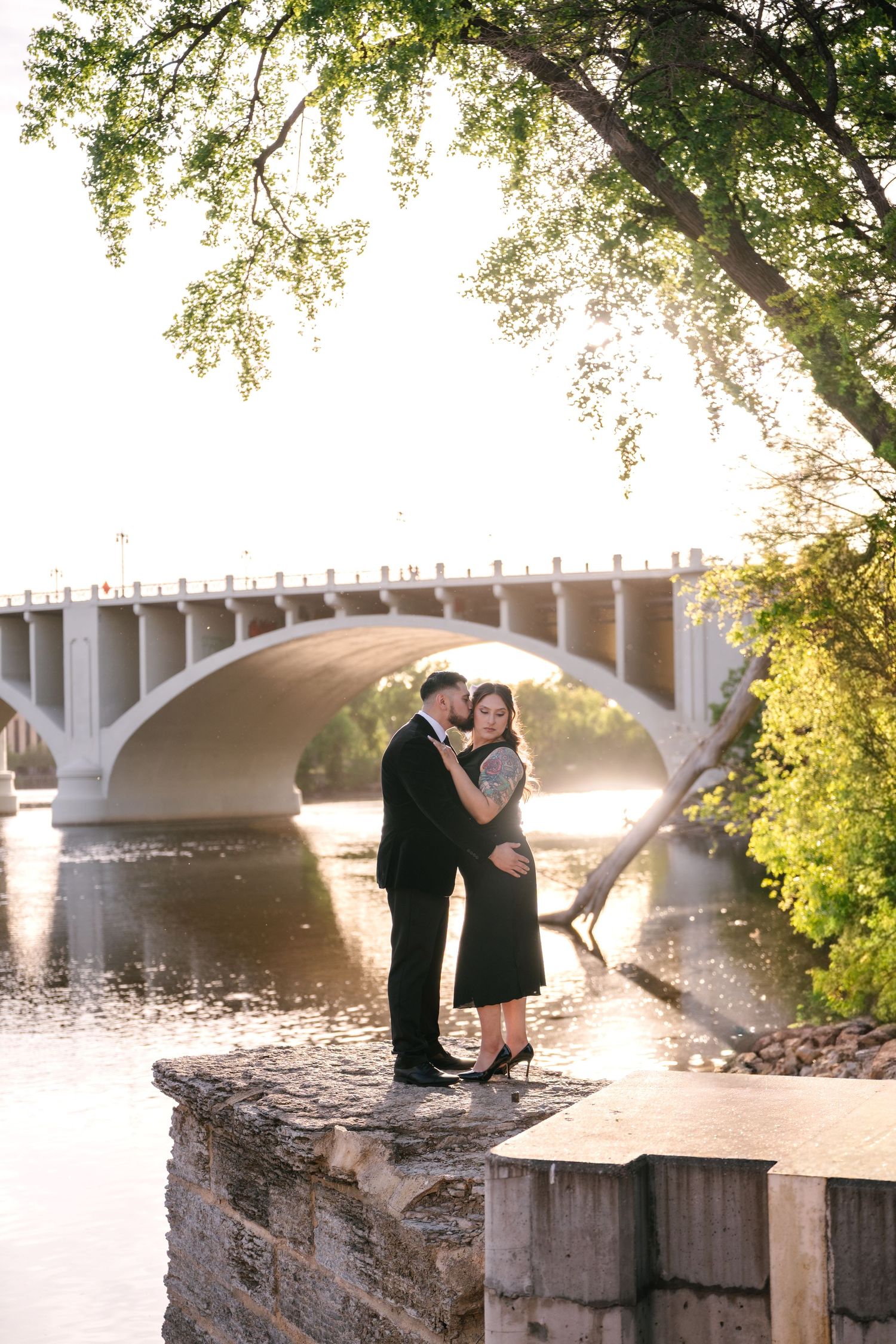 Romantic couple embracing on stone wall with arched bridge and river at golden hour.
