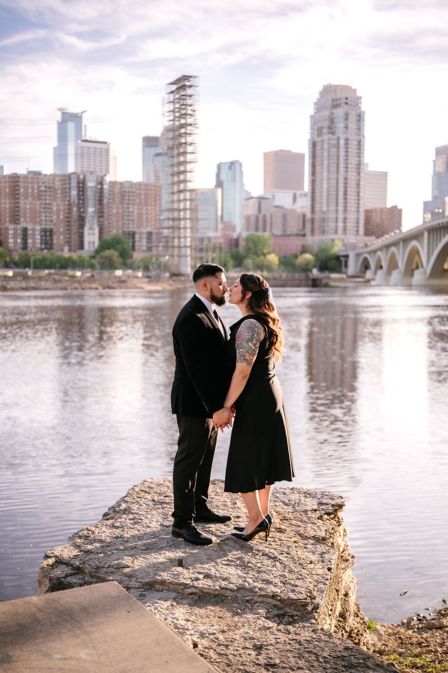 Couple kissing on rocky riverbank with Minneapolis city skyline reflected in water.