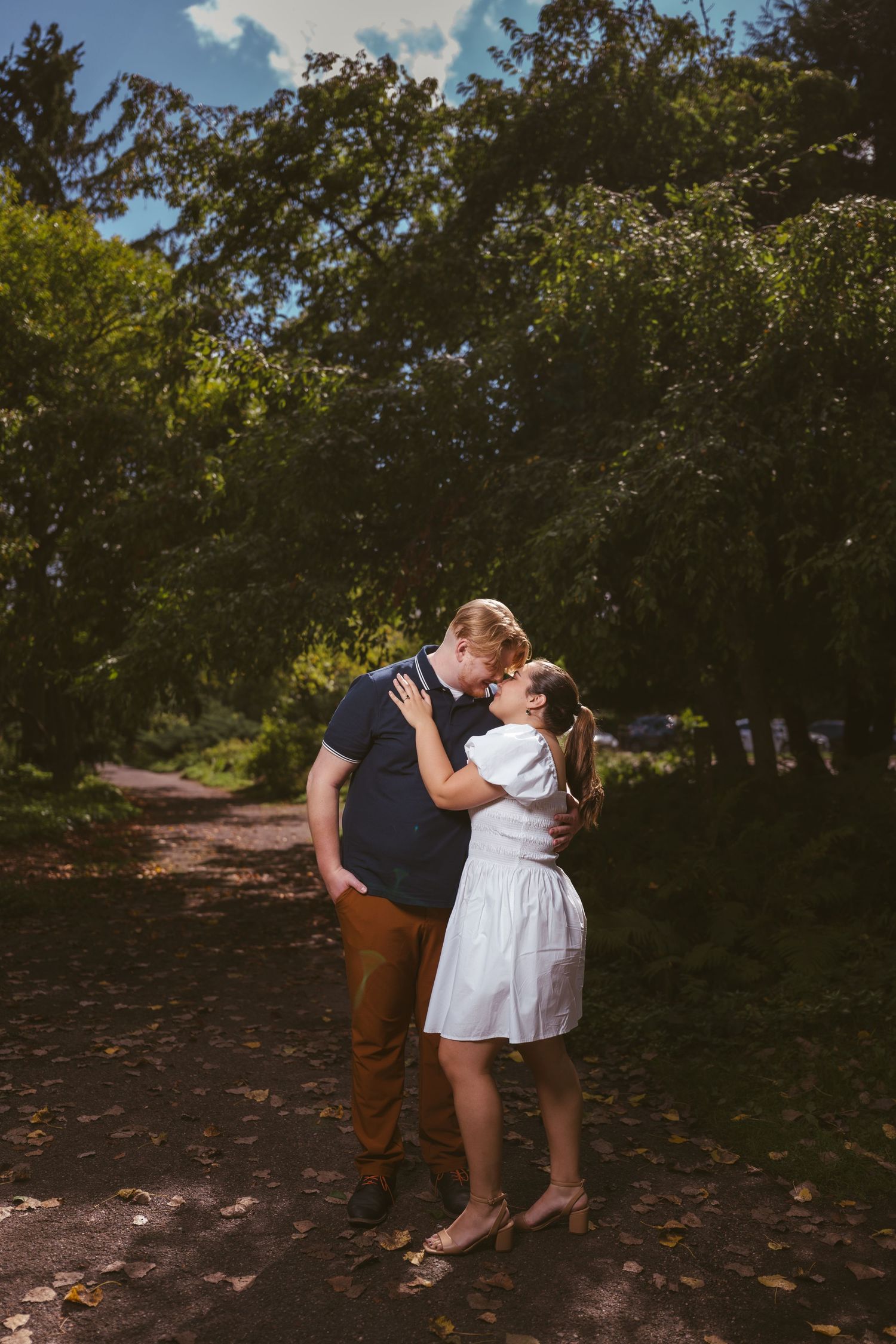 Couple shares romantic moment on tree-lined path in dappled sunlight.