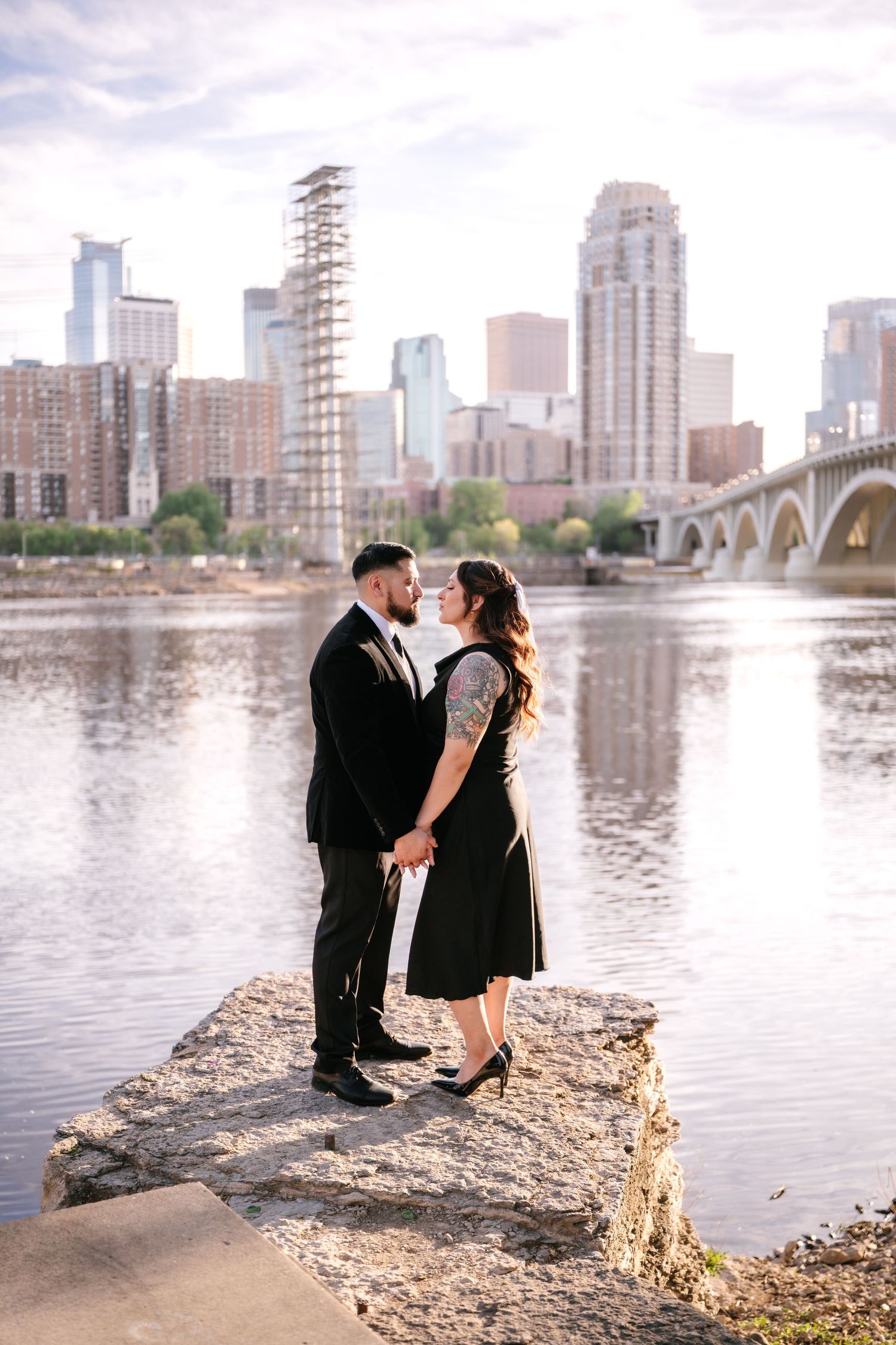 Couple holding hands on rocky riverbank with Minneapolis skyline and bridge reflecting in the water.