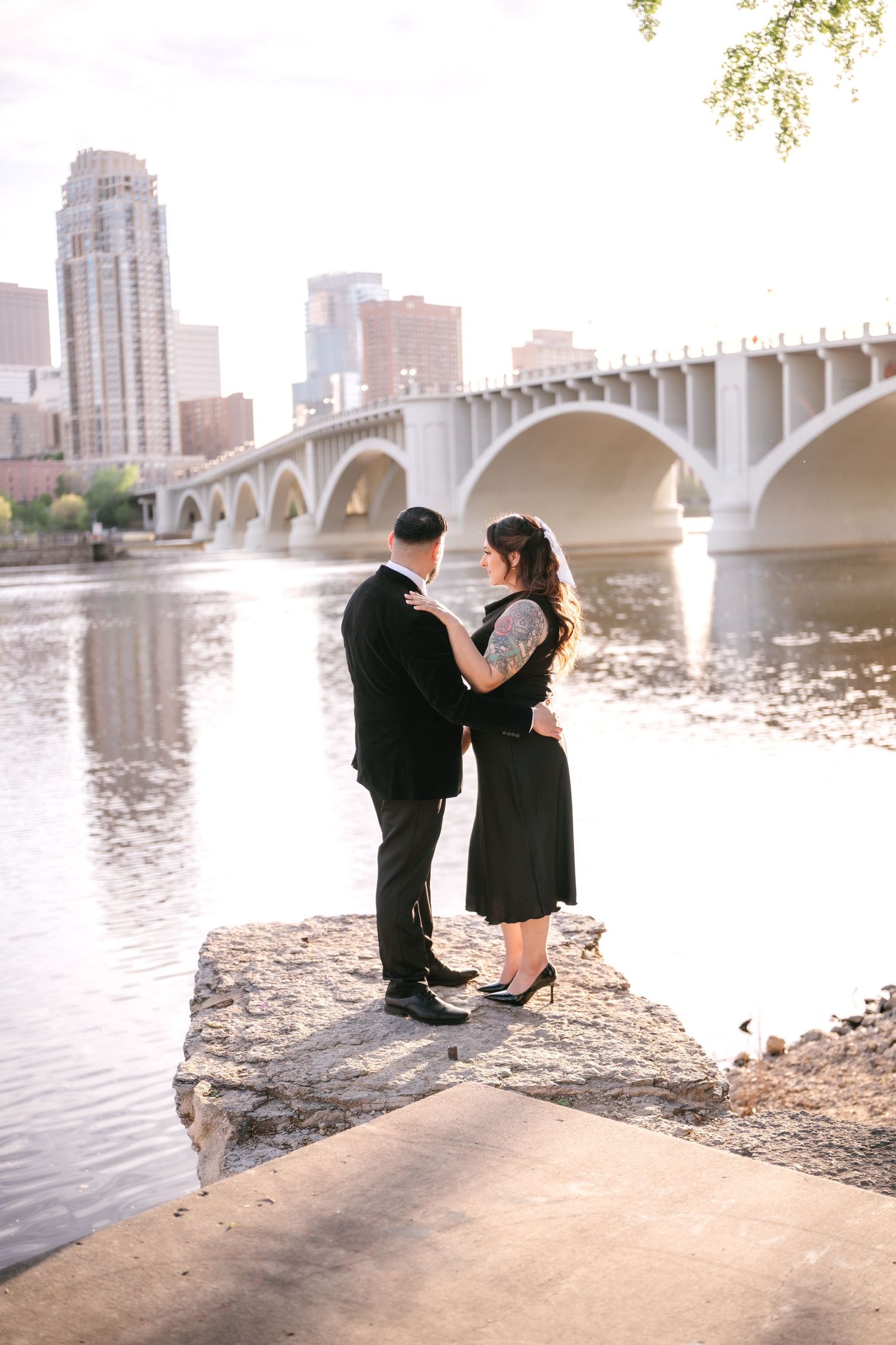 Couple stands on rocky riverbank with city bridge and skyline backdrop during golden hour engagement session.