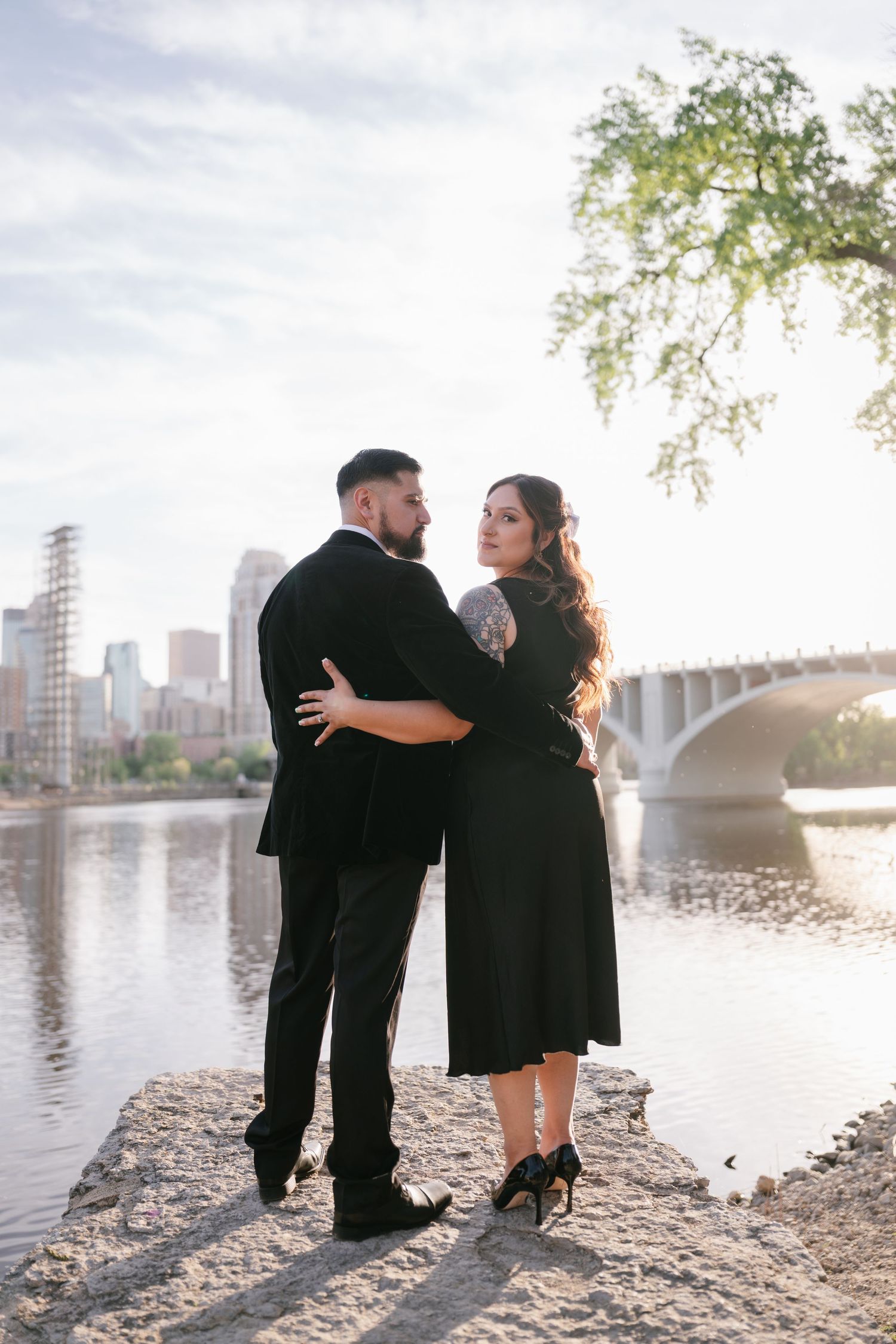 Couple in black formal wear posing by riverside with city bridge in background.