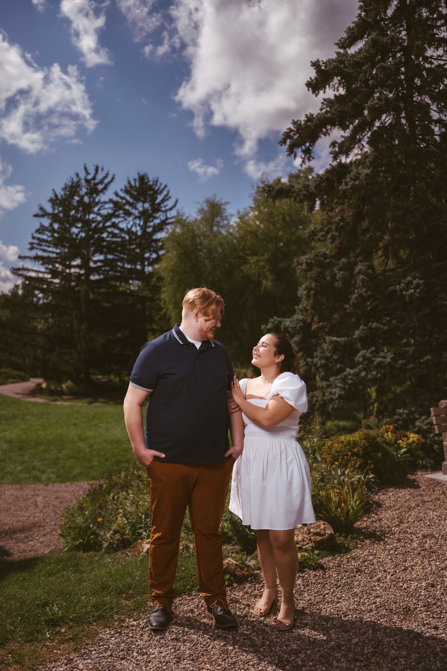 A romantic couple posing together on a gravel path surrounded by evergreen trees.