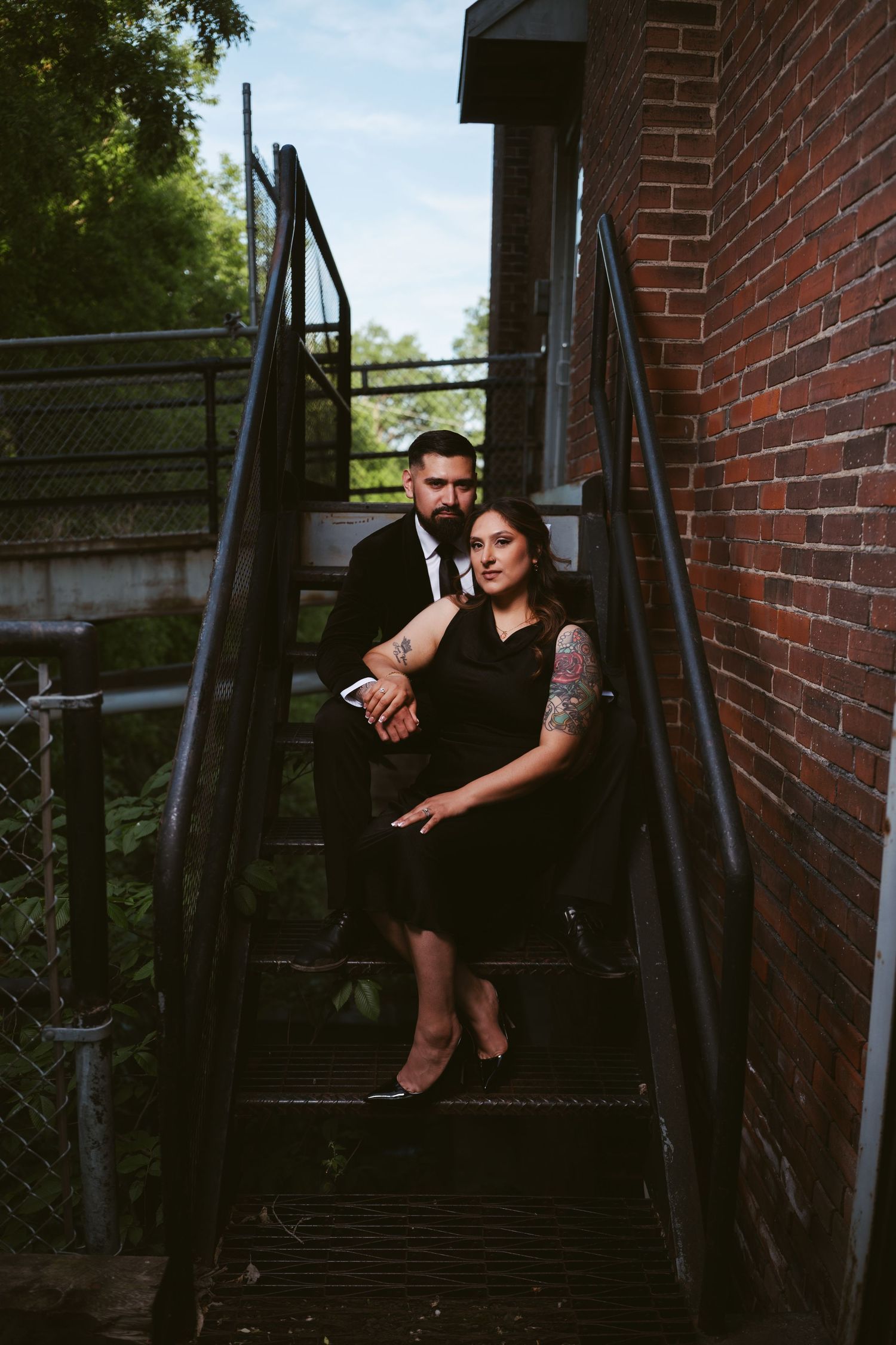 Elegant couple in black posing on industrial fire escape stairs beside a brick wall.