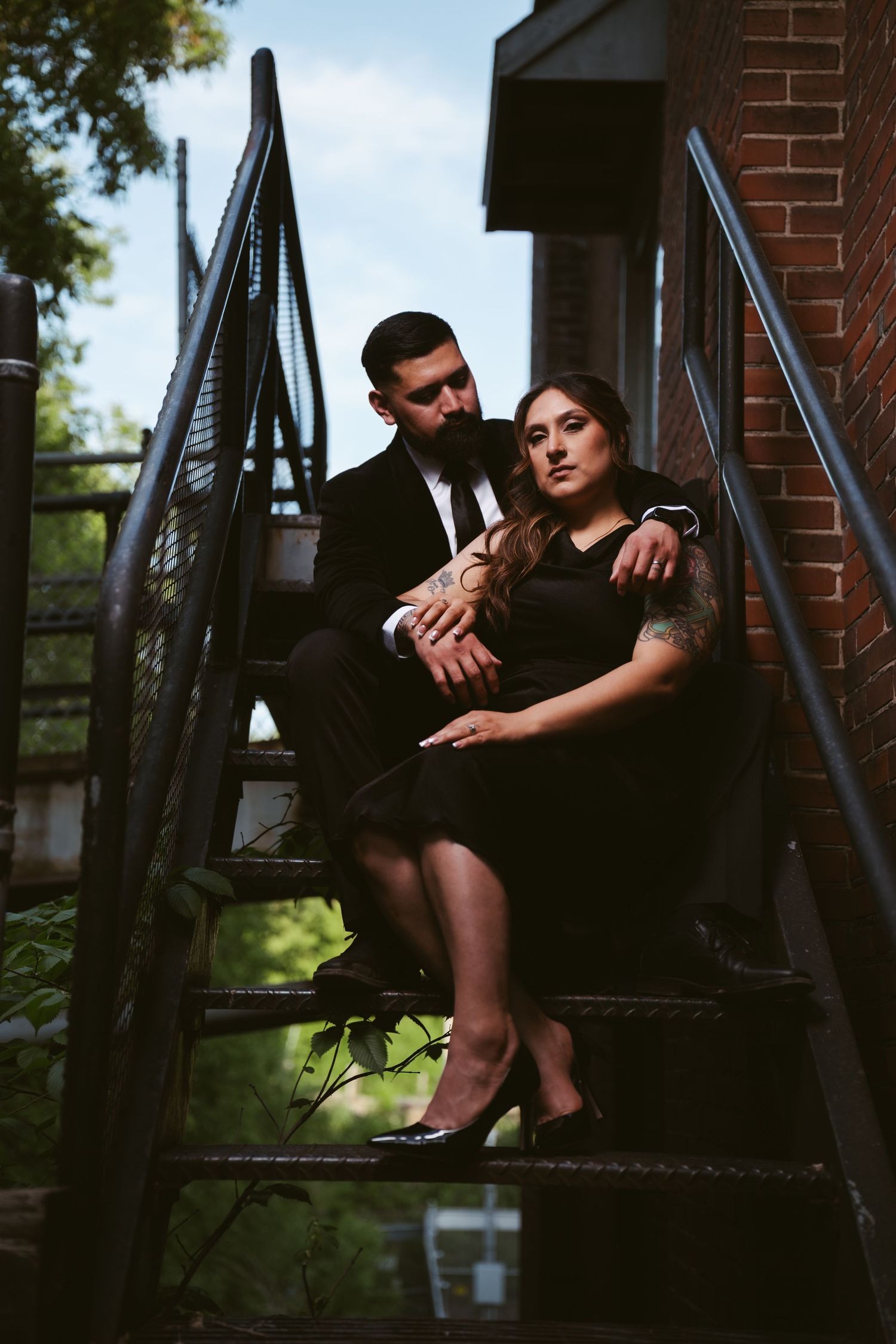A couple in black formal attire sits closely on metal fire escape stairs beside a brick building.