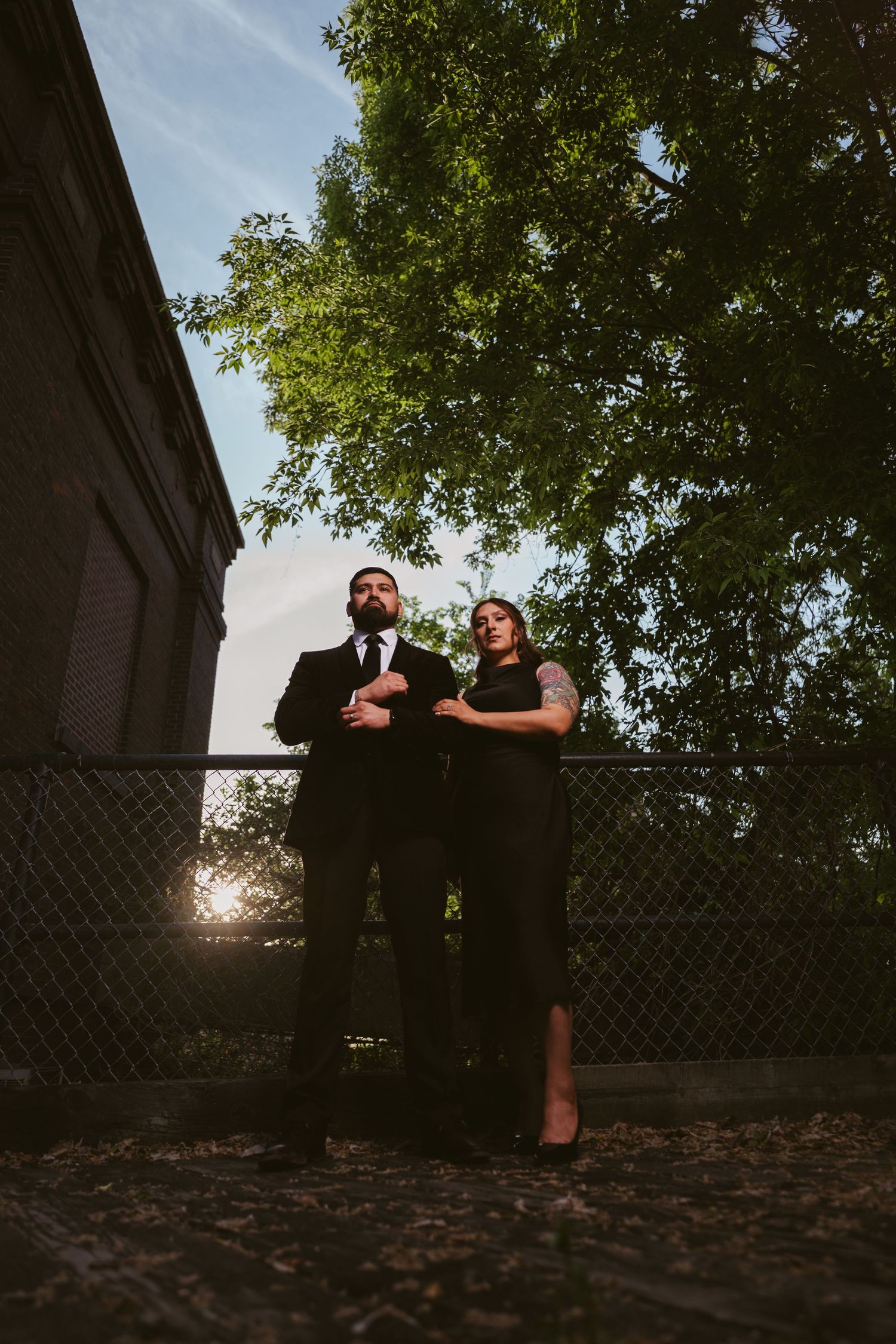 Couple in black formal attire standing by a chain-link fence with lush green trees and golden sunlight behind them.