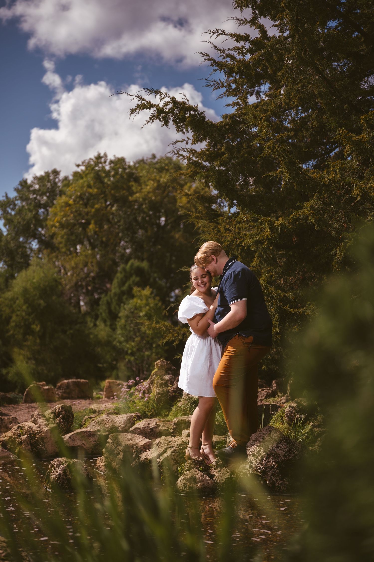 A couple sharing an intimate moment near a pond with rocks and trees in the background.