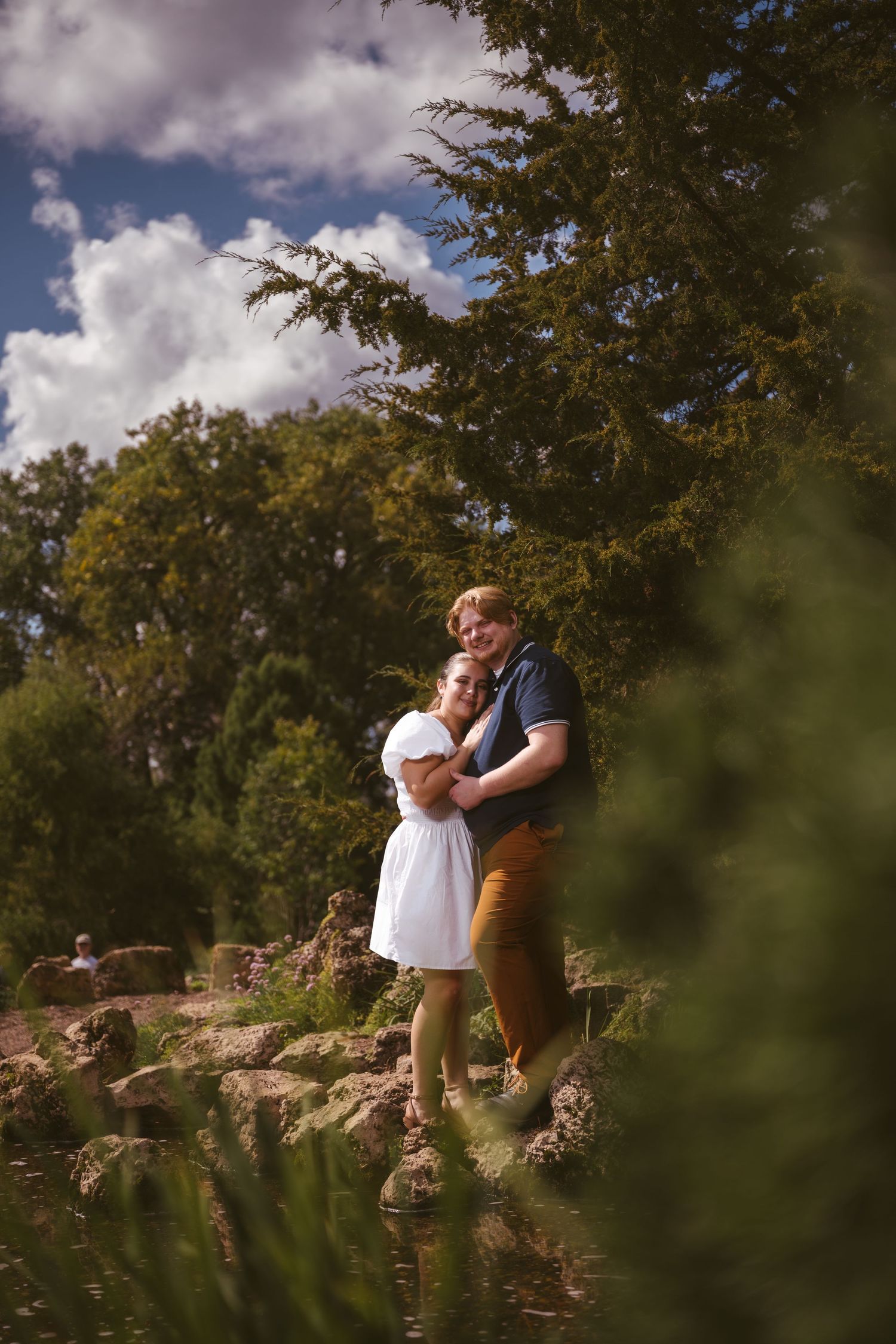 A couple embracing on rocky landscaping beside a tranquil pond.