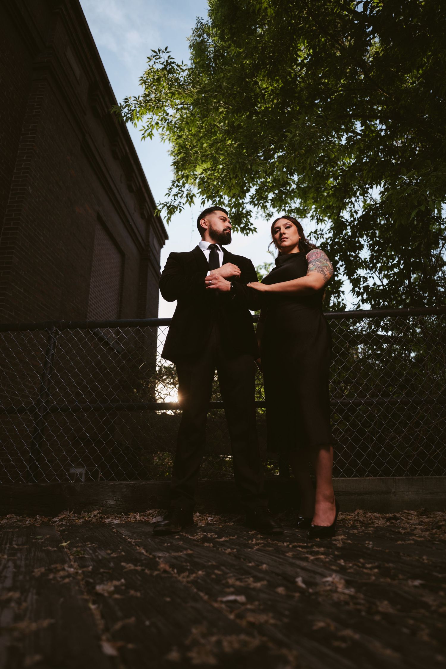 Stylish couple in black formal wear posing dramatically under trees near a brick building at dusk.