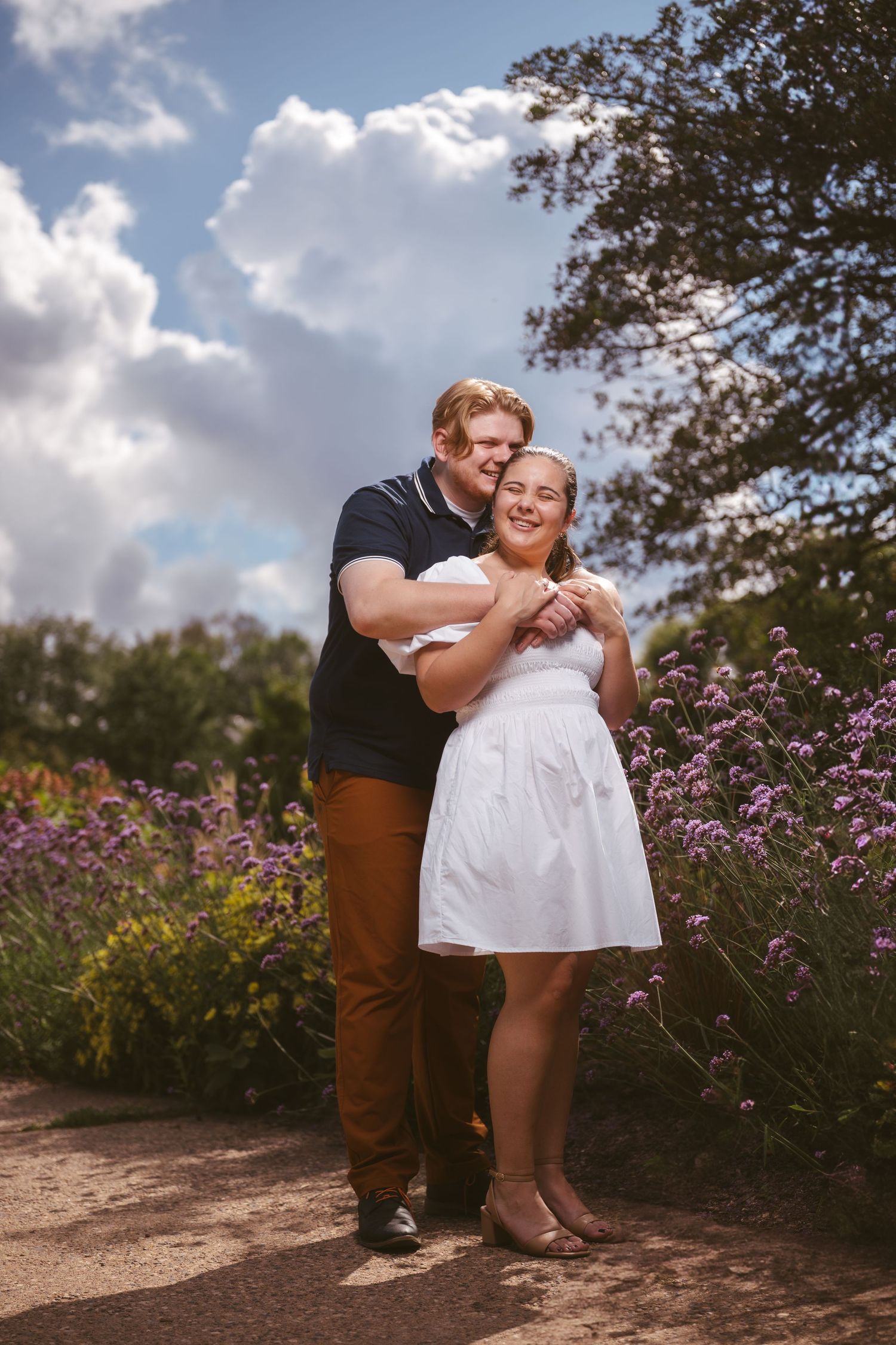 A romantic couple embracing near blooming purple flowers on a sunny afternoon.