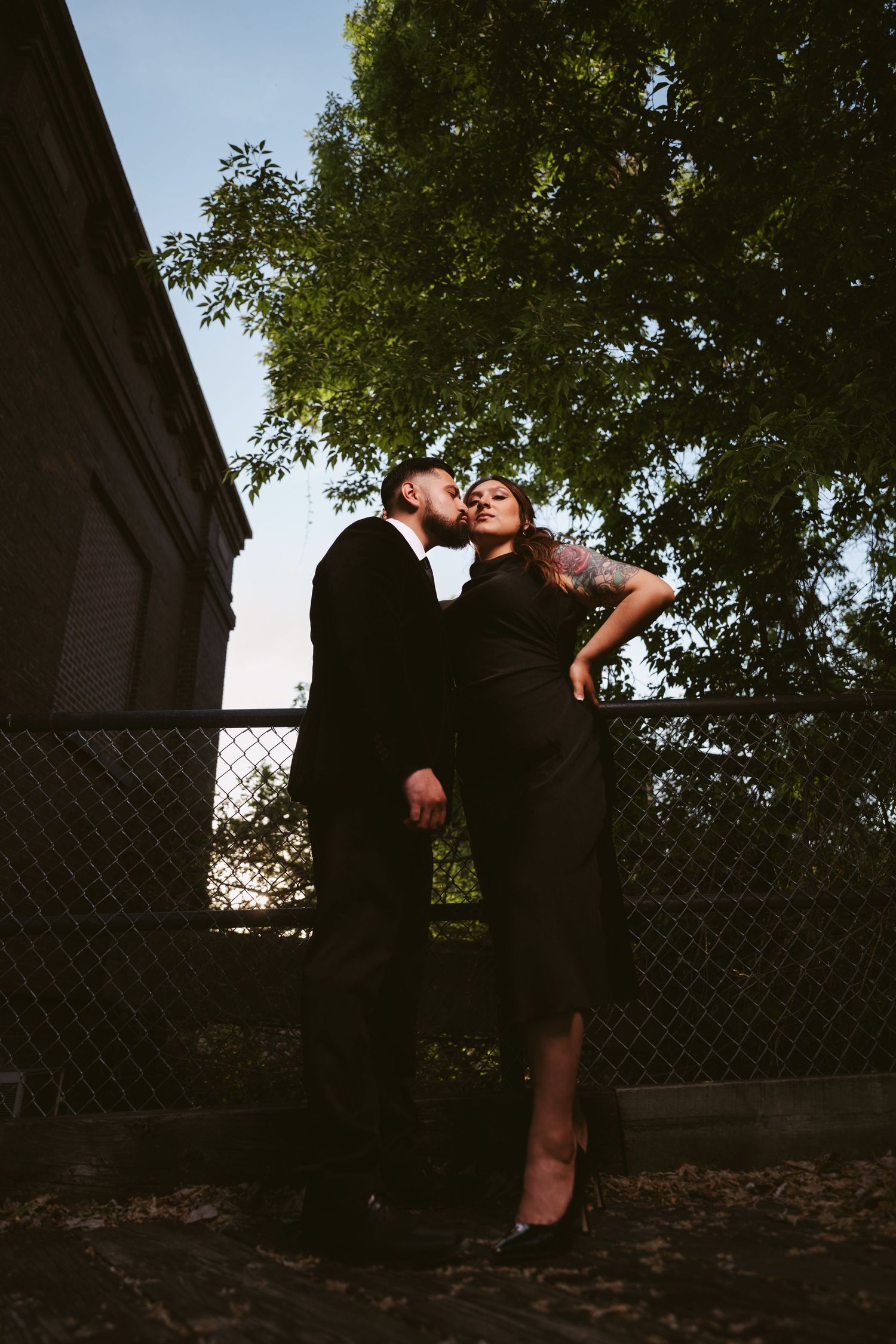Couple in matching black outfits sharing a tender kiss outdoors near a fence with lush green trees.