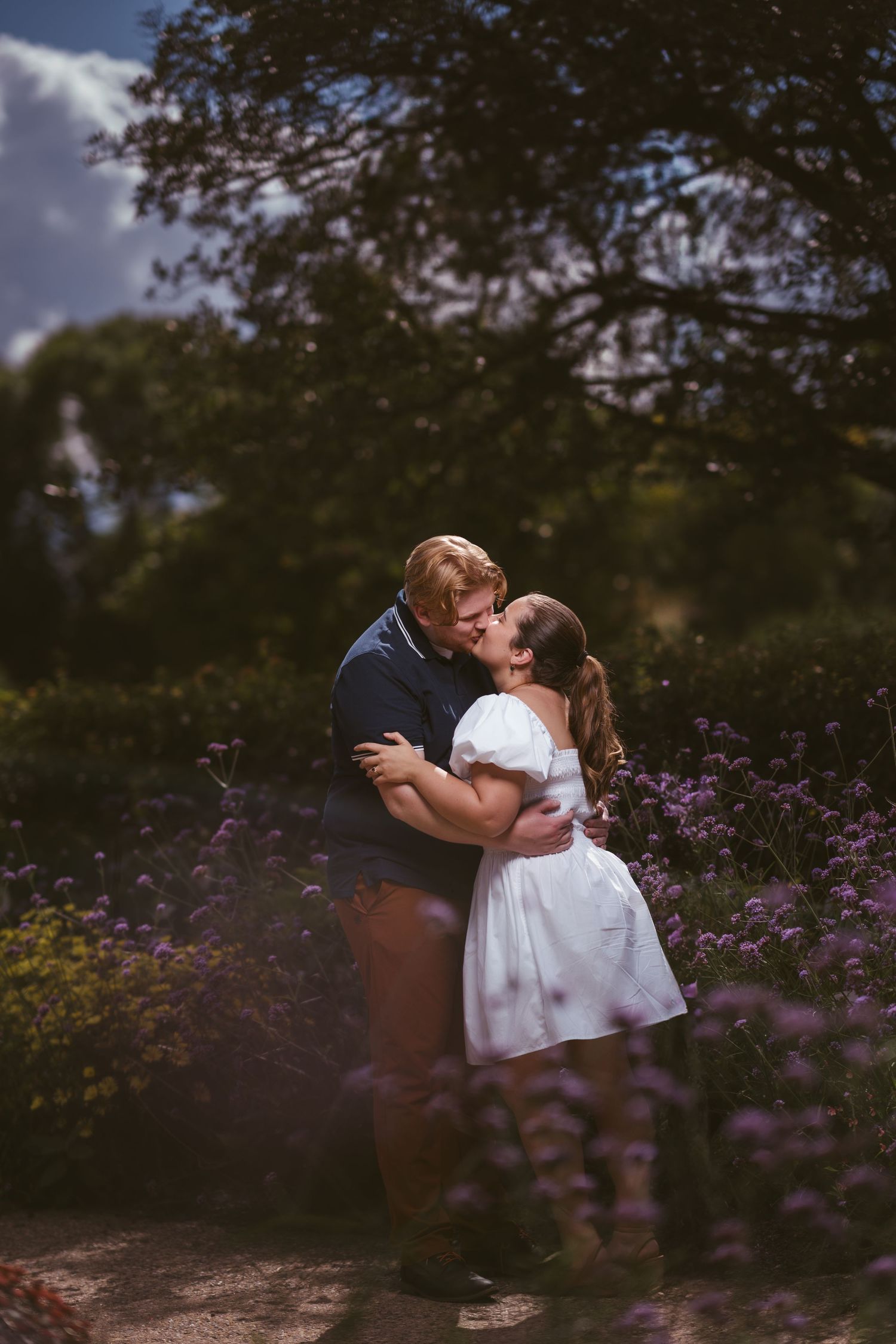 A couple sharing an intimate kiss surrounded by purple wildflowers and natural scenery.