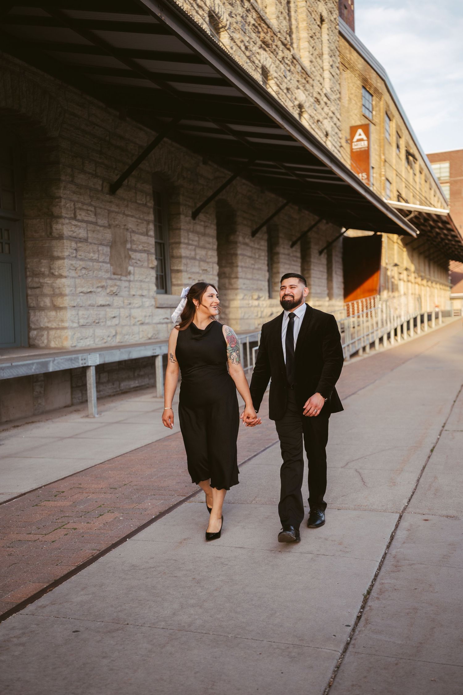 Stylish couple holding hands walking past historic stone building at sunset.