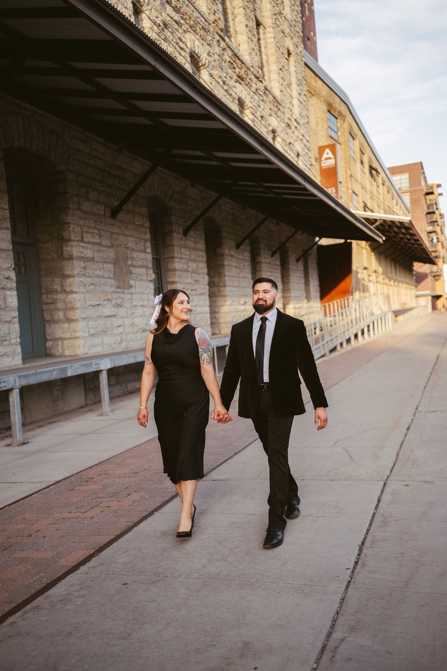 Stylish couple in black outfits walk hand in hand along a sunny brick walkway beside a historic building.