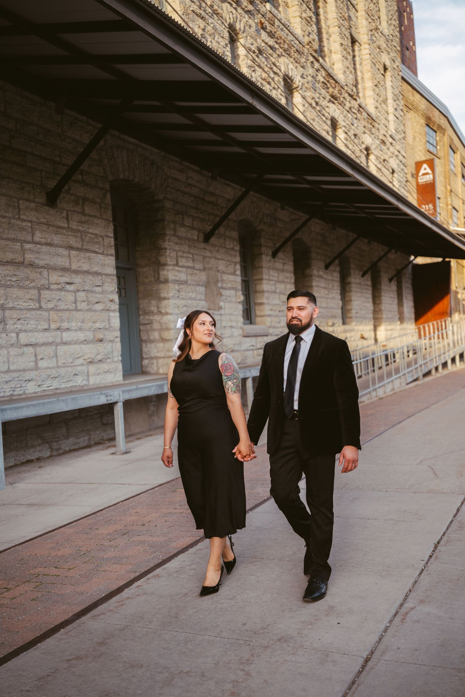 Couple holding hands walking past historic stone building in matching black outfits.