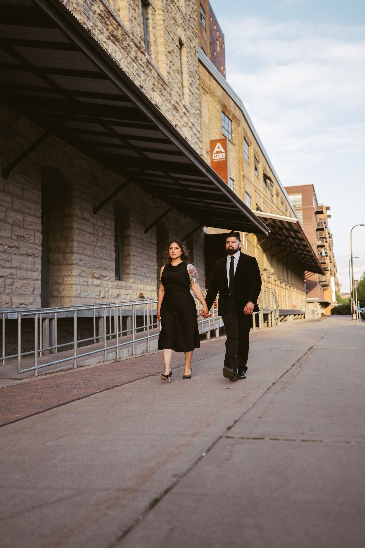 Couple in black formal attire strolling along sidewalk beside stone building.