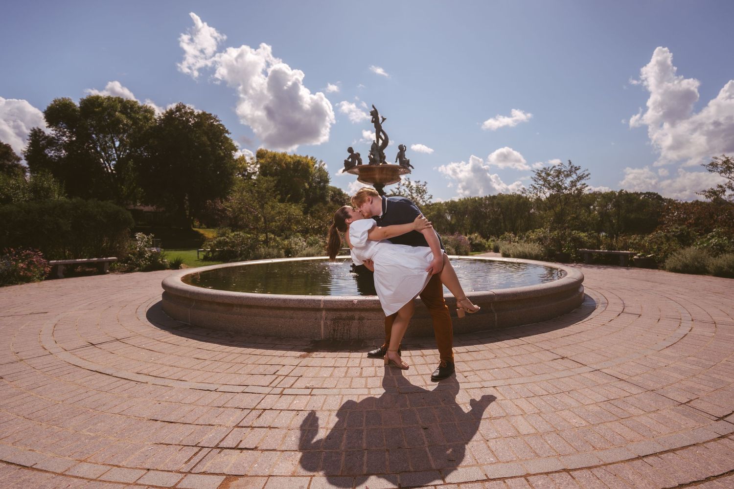 A couple stands together near a circular stone fountain in a manicured garden.