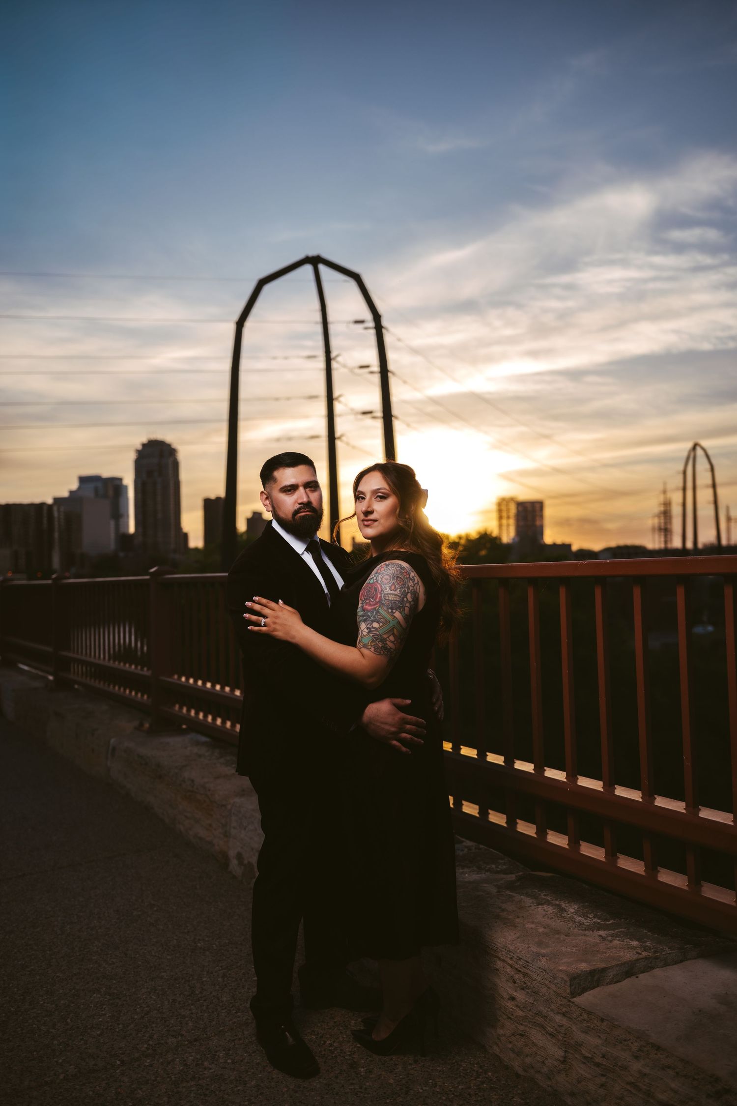 Couple embracing on a bridge at sunset with city skyline backdrop, woman with tattoos in black dress, man in suit.