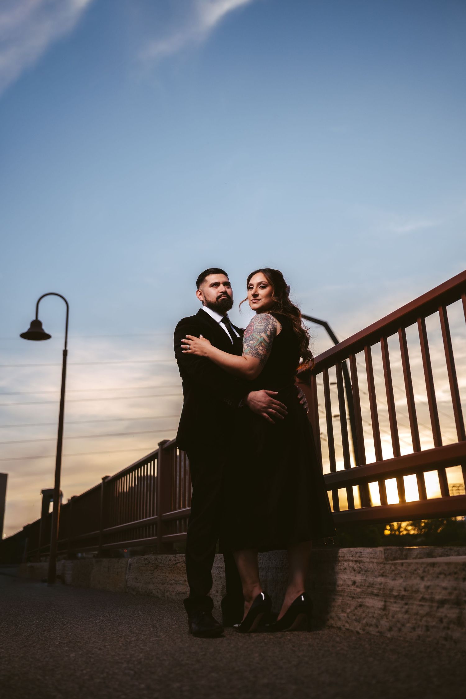 Couple embracing at sunset on a bridge with warm sky and city lights behind them.