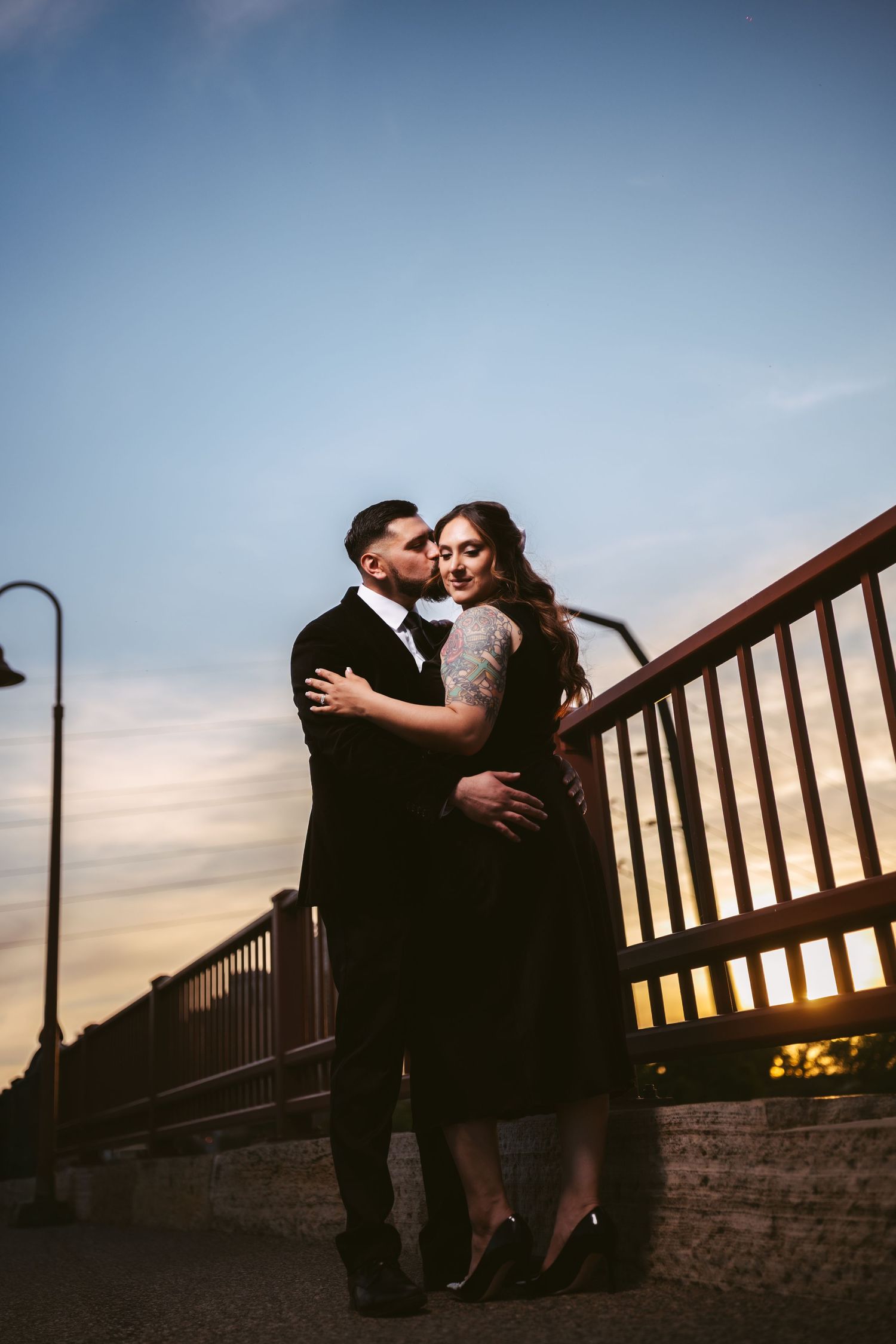 Couple embracing romantically on a bridge at sunset, man in black suit and woman in black dress with tattoos.