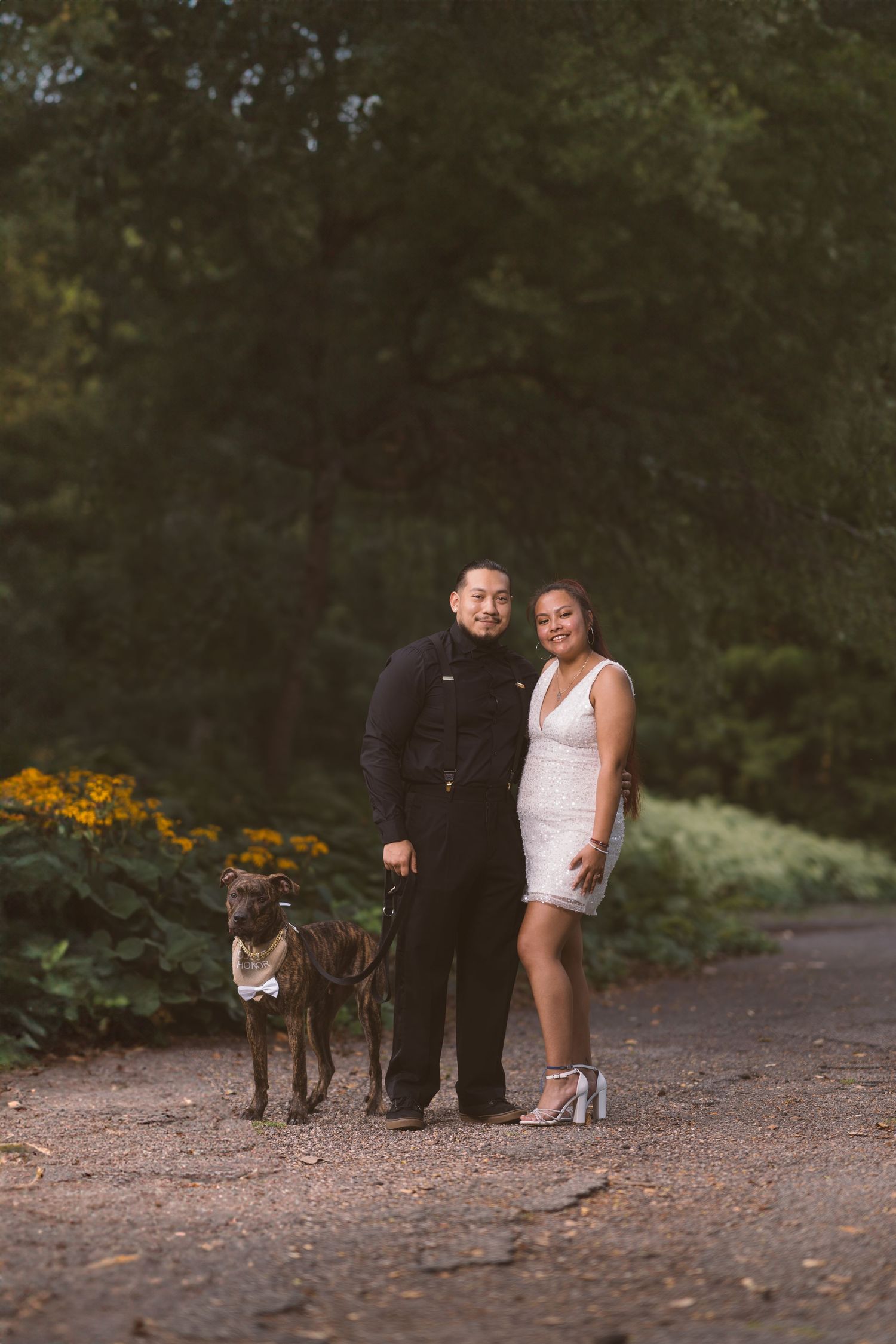 A couple poses with their dog on a garden path surrounded by yellow flowers and greenery.