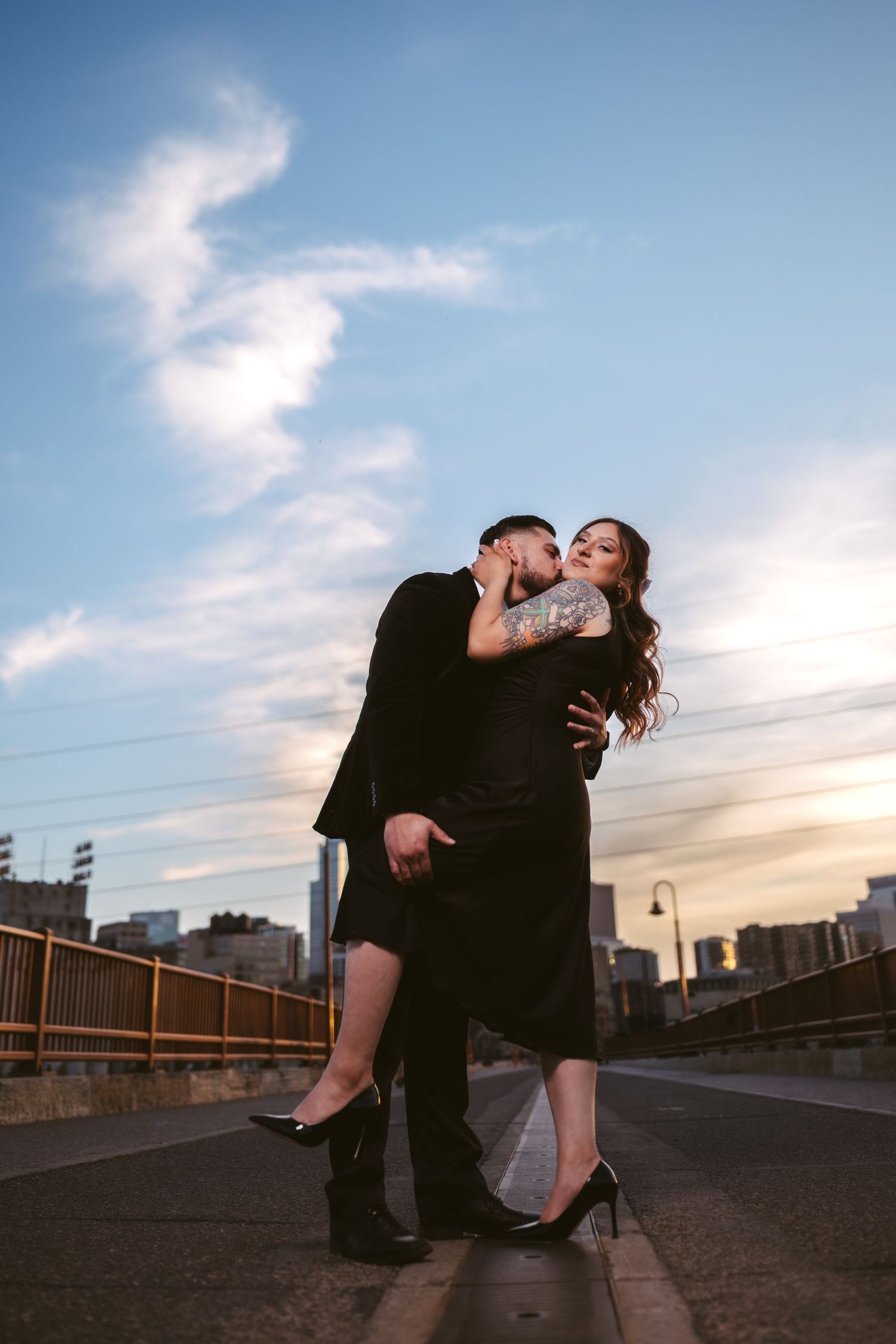 Couple sharing a passionate kiss on urban bridge at sunset with city skyline.