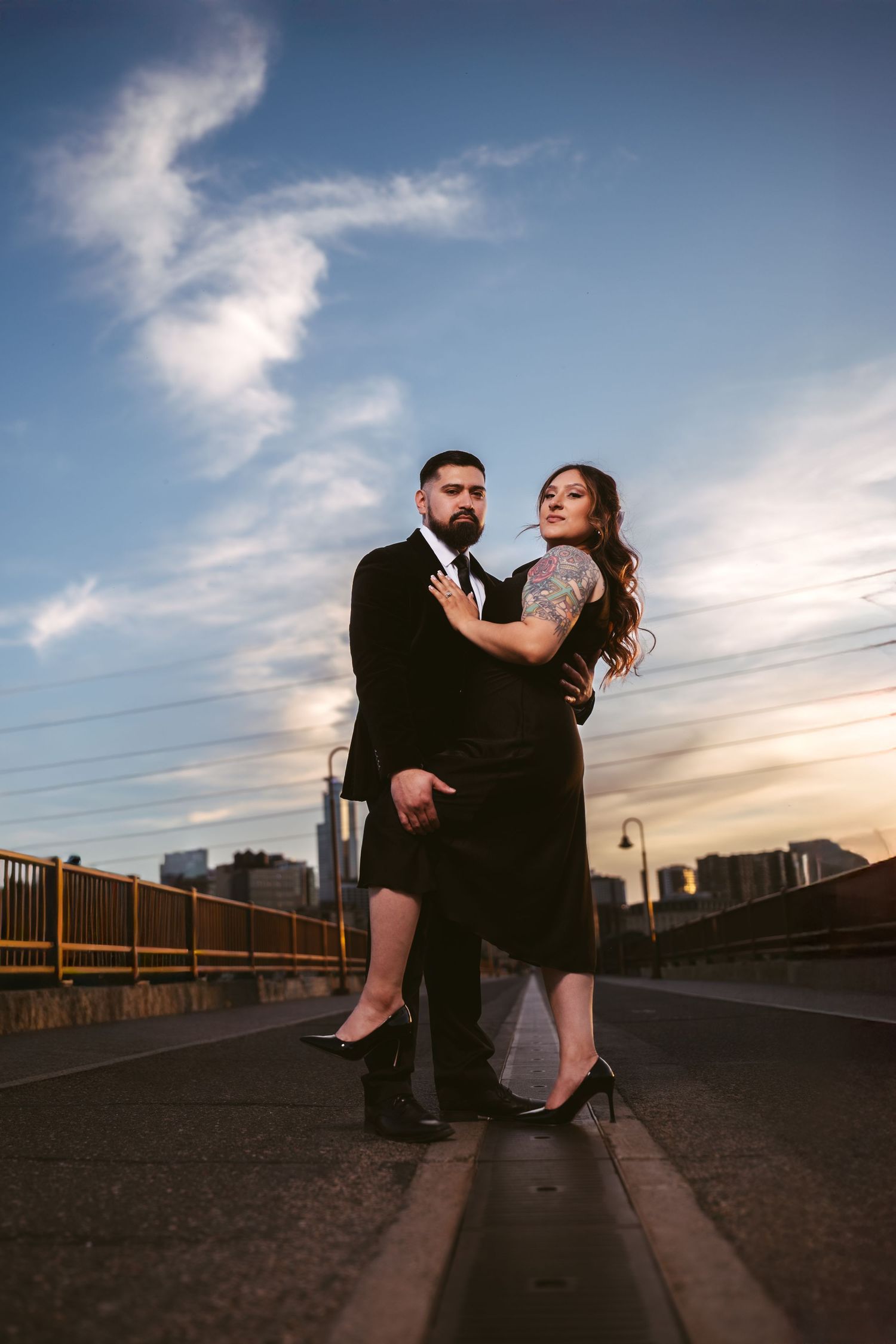 Elegant couple in black formal attire posing on a bridge at sunset with city skyline in background.