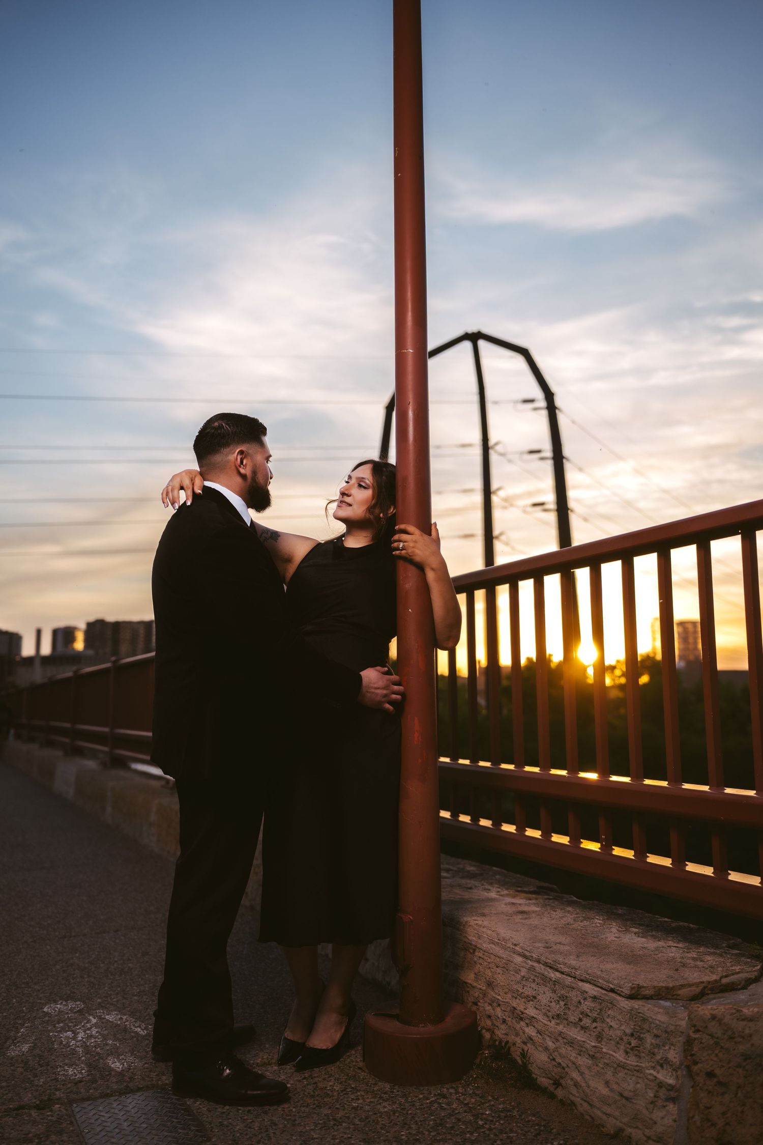 Couple in black formal wear embracing by a lamp post on a bridge during a vibrant golden sunset.
