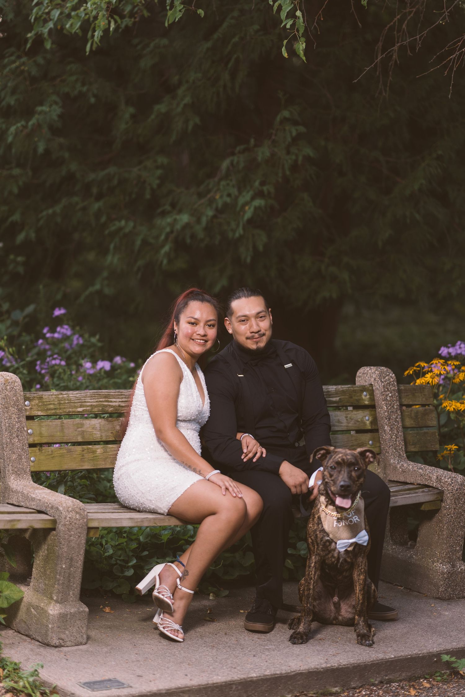 A couple sits on a wooden bench in a garden with their dog beside them.