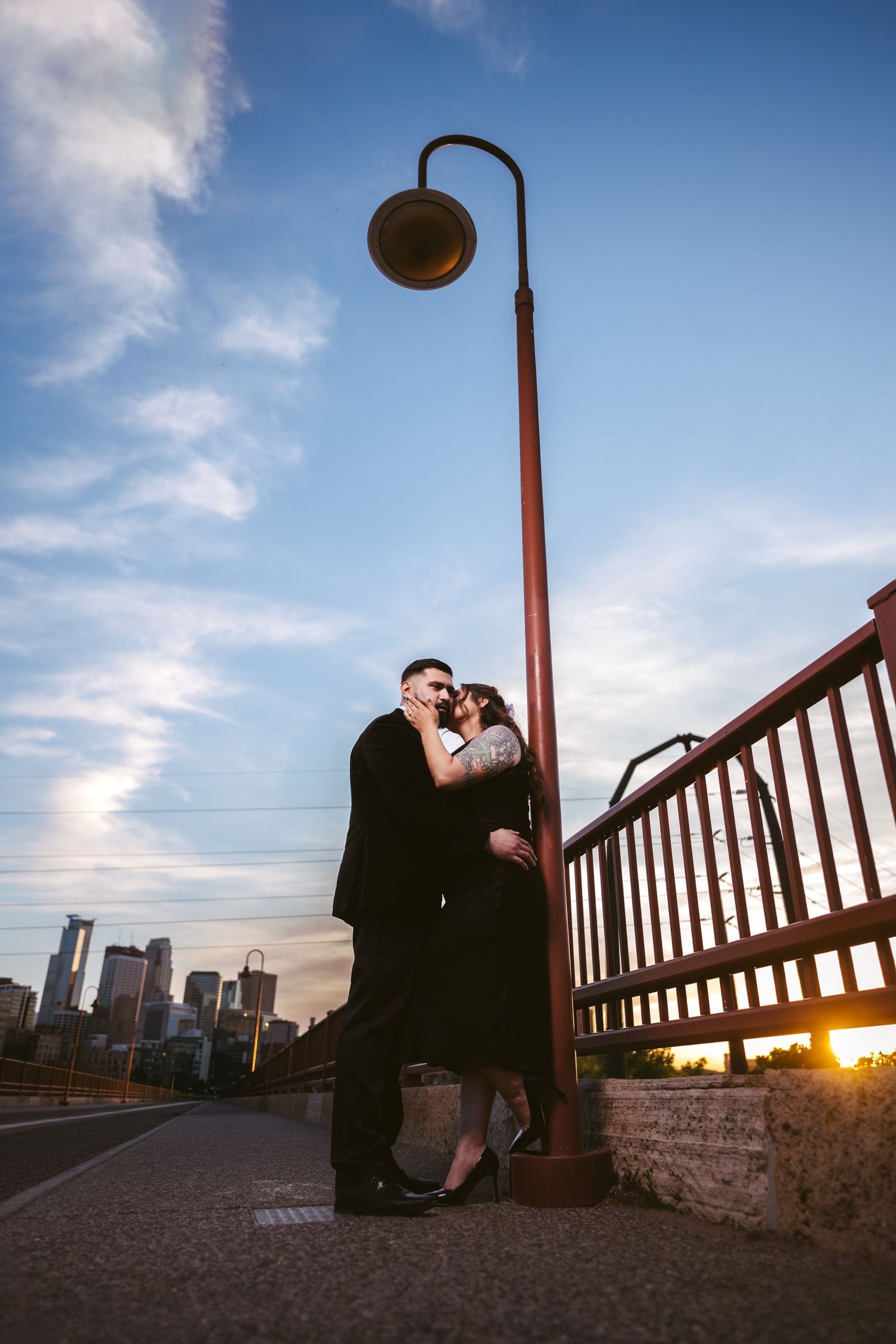 Couple kissing by a vintage street lamp on a bridge at sunset with city skyline in the background.