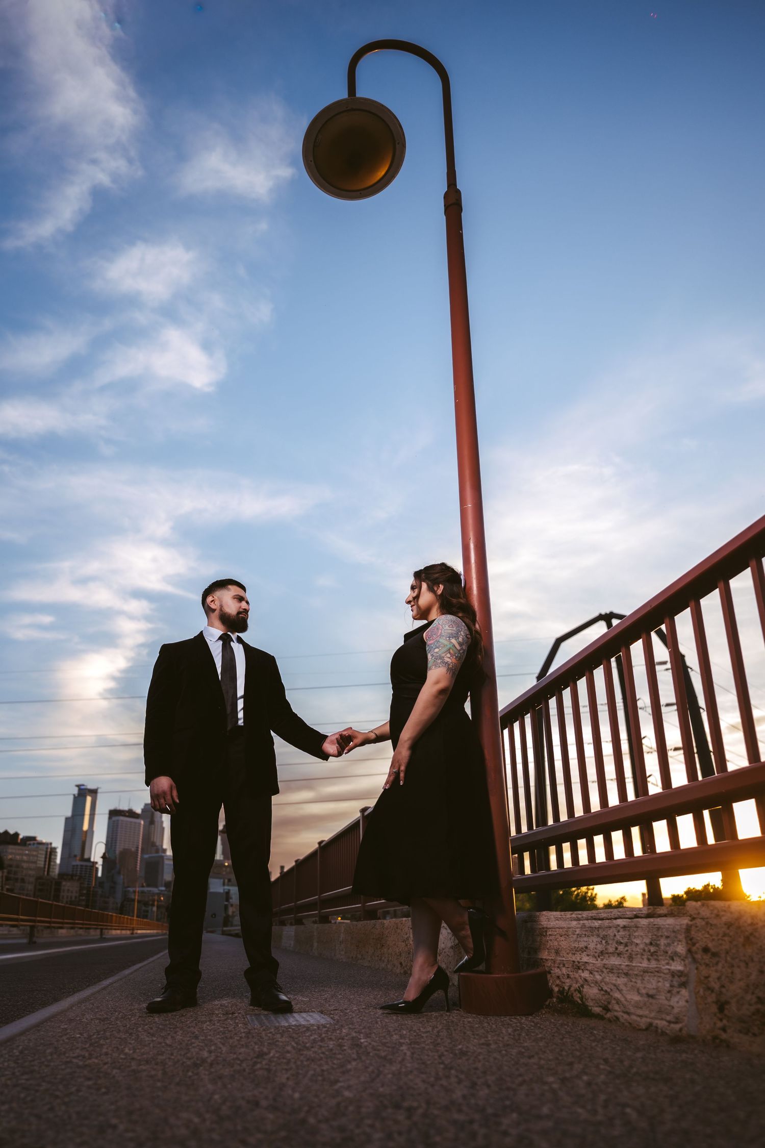 Couple holds hands under vintage street lamp on bridge at sunset with city skyline in background.
