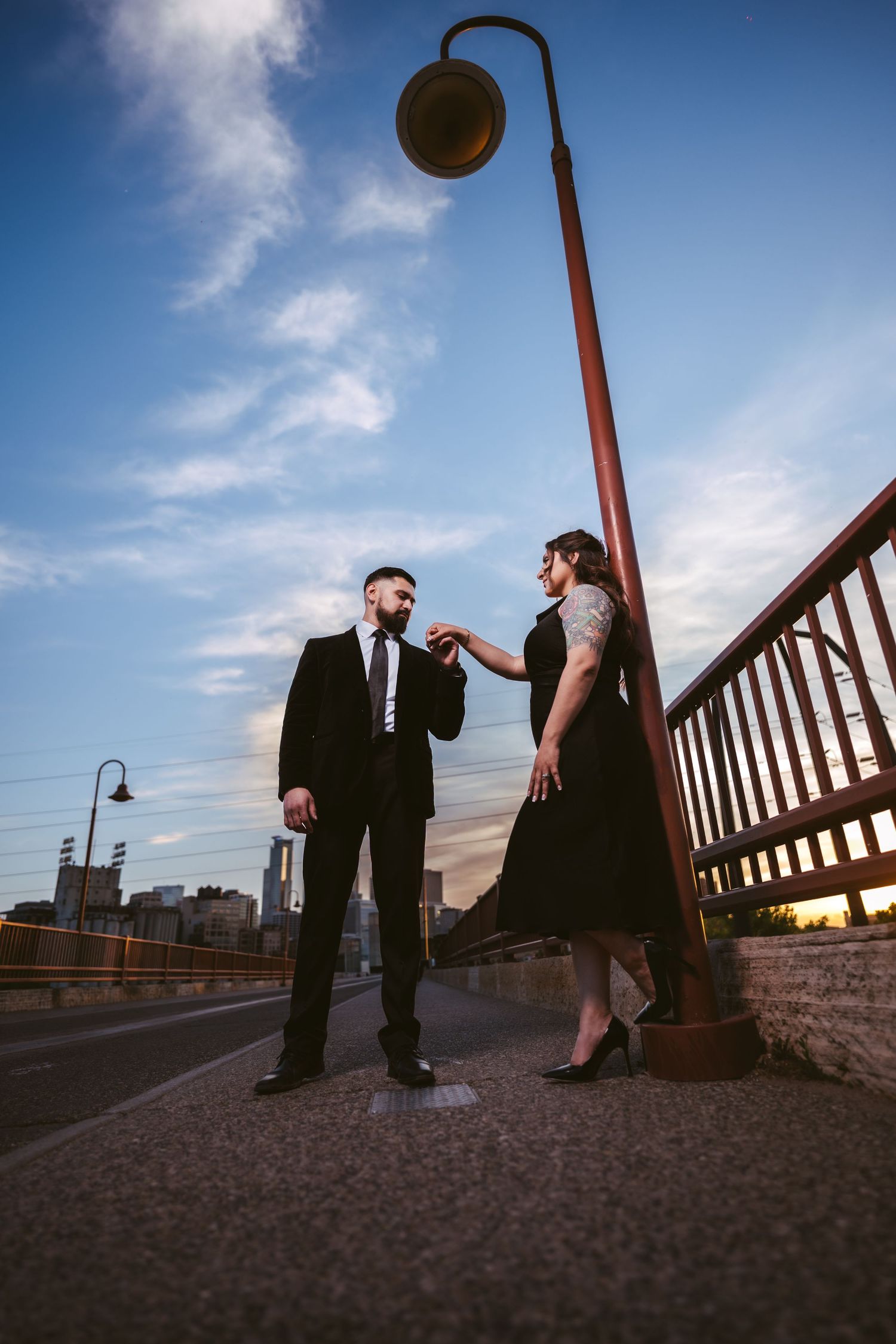 Couple in black formal wear share a tender moment by a lamppost on a bridge at sunset.