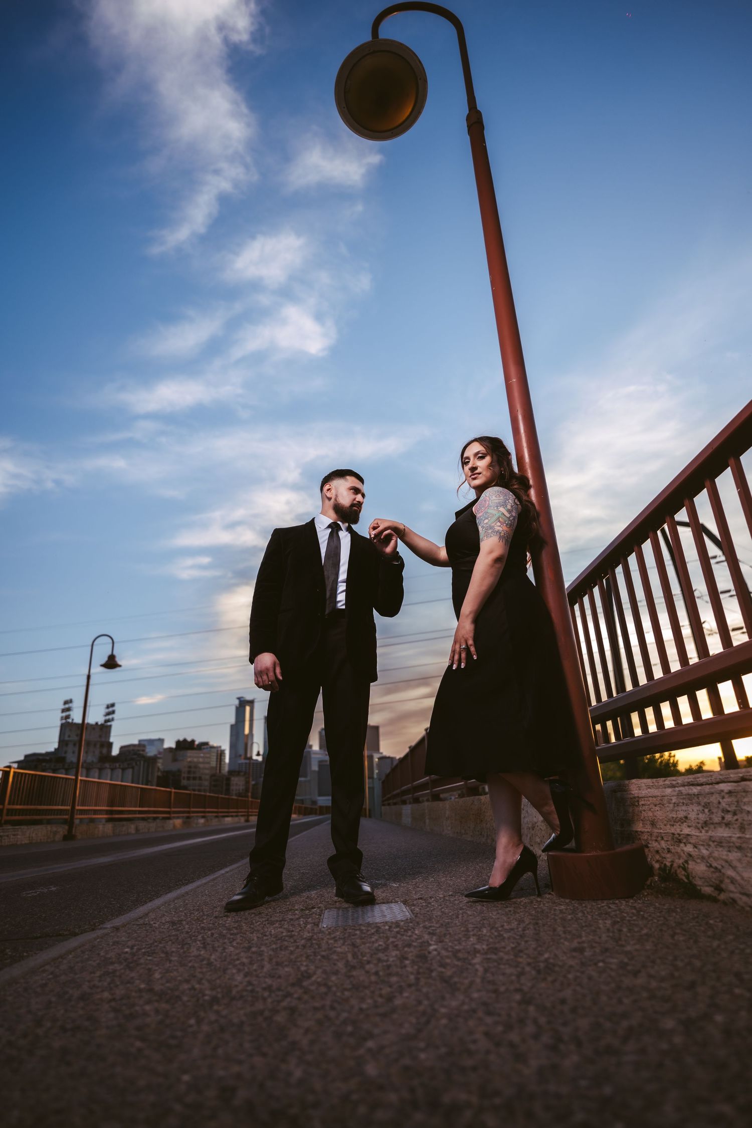 Couple posing under a street lamp on a bridge at dusk, man in black suit and woman in black dress.