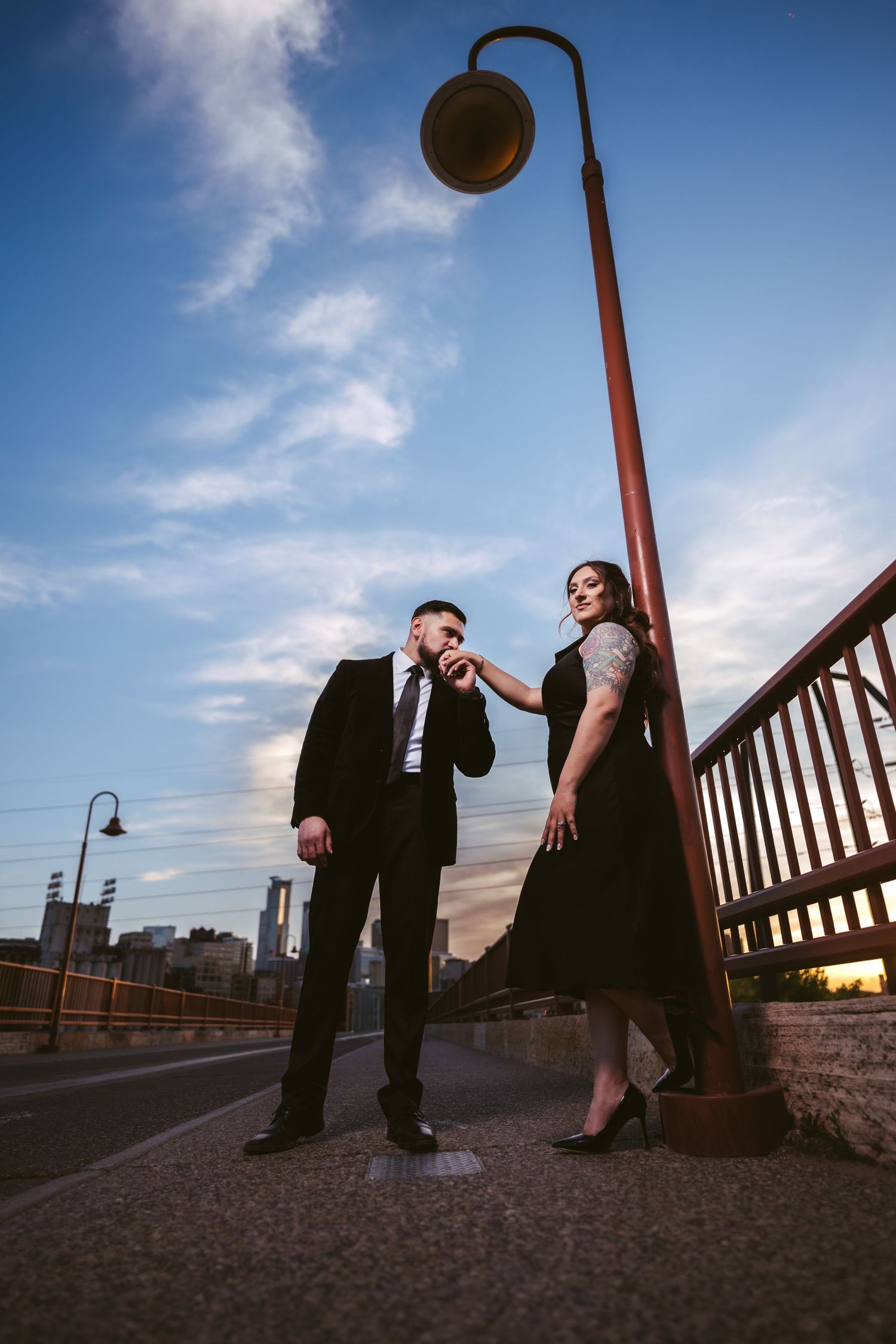 Man kisses womans hand by a red lamppost on a bridge at dusk with city skyline in background.