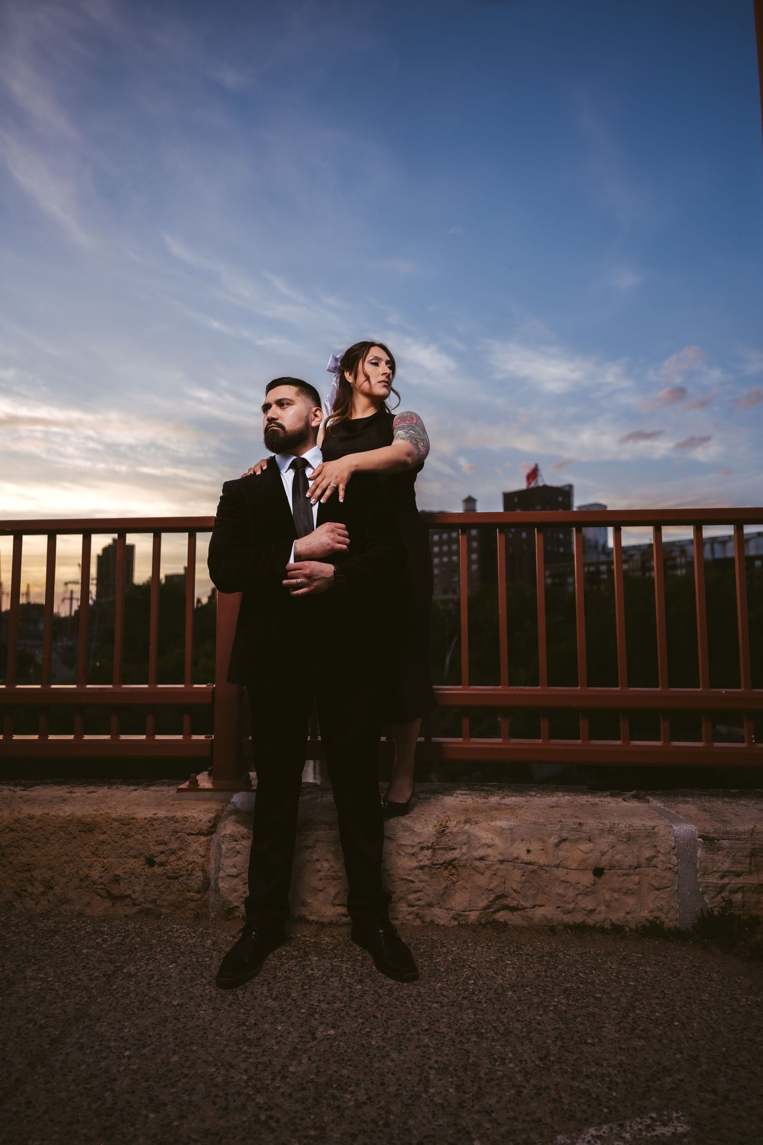 Couple in black formal attire standing on a stone ledge by a bridge railing at colorful sunset.