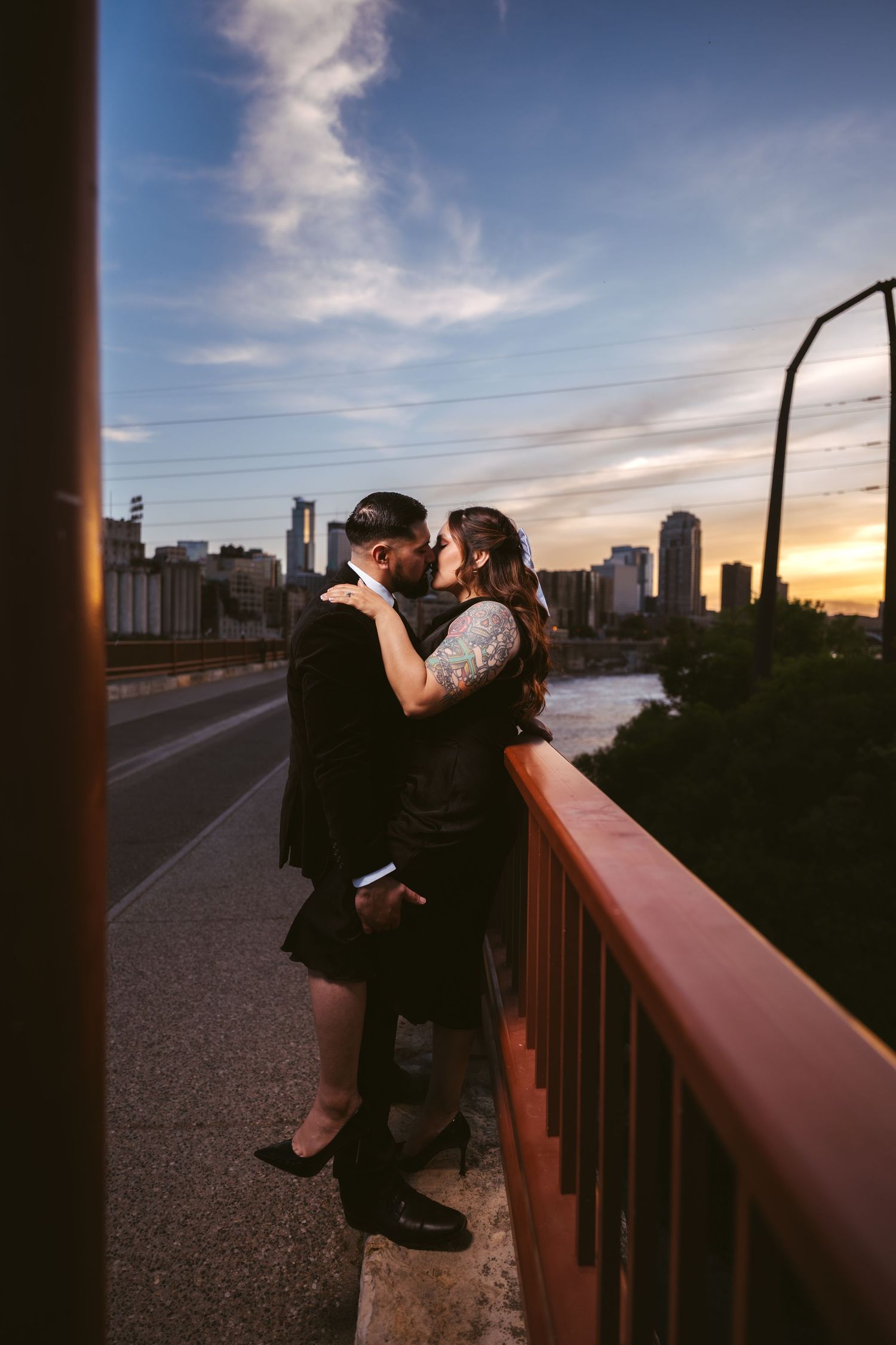 Couple embracing on bridge at sunset with city skyline glowing in background.