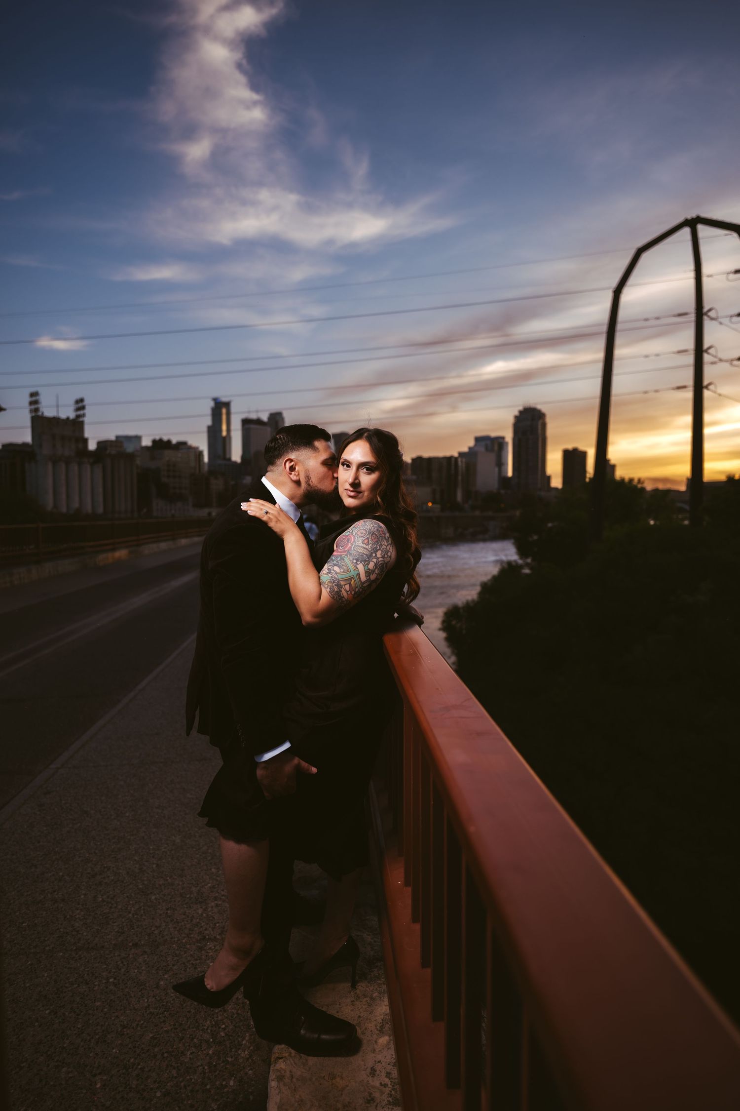 Couple cuddling on bridge railing at dusk with urban skyline behind them.