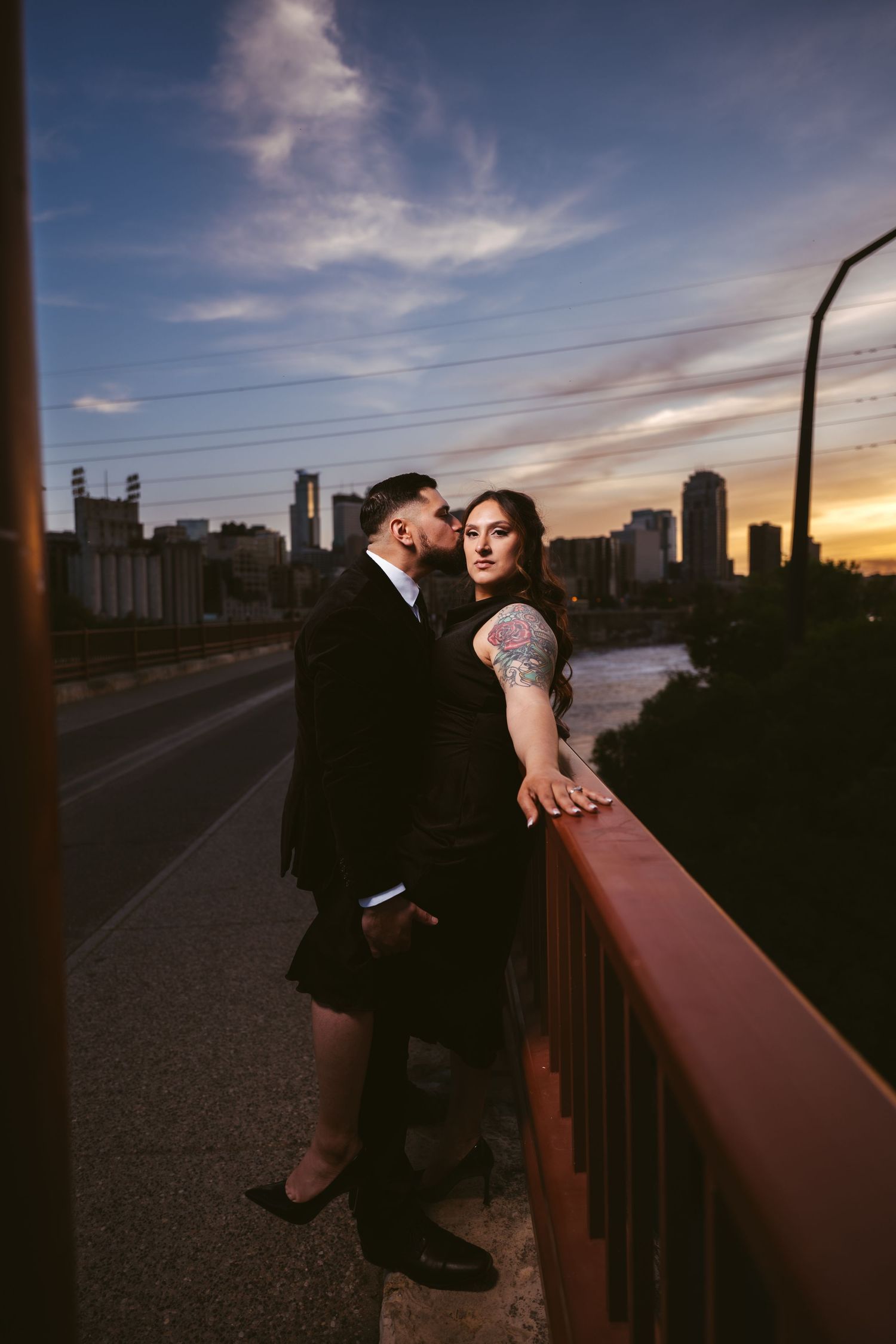 Couple embraces on urban bridge railing at dusk with colorful sunset sky and city skyline behind them.
