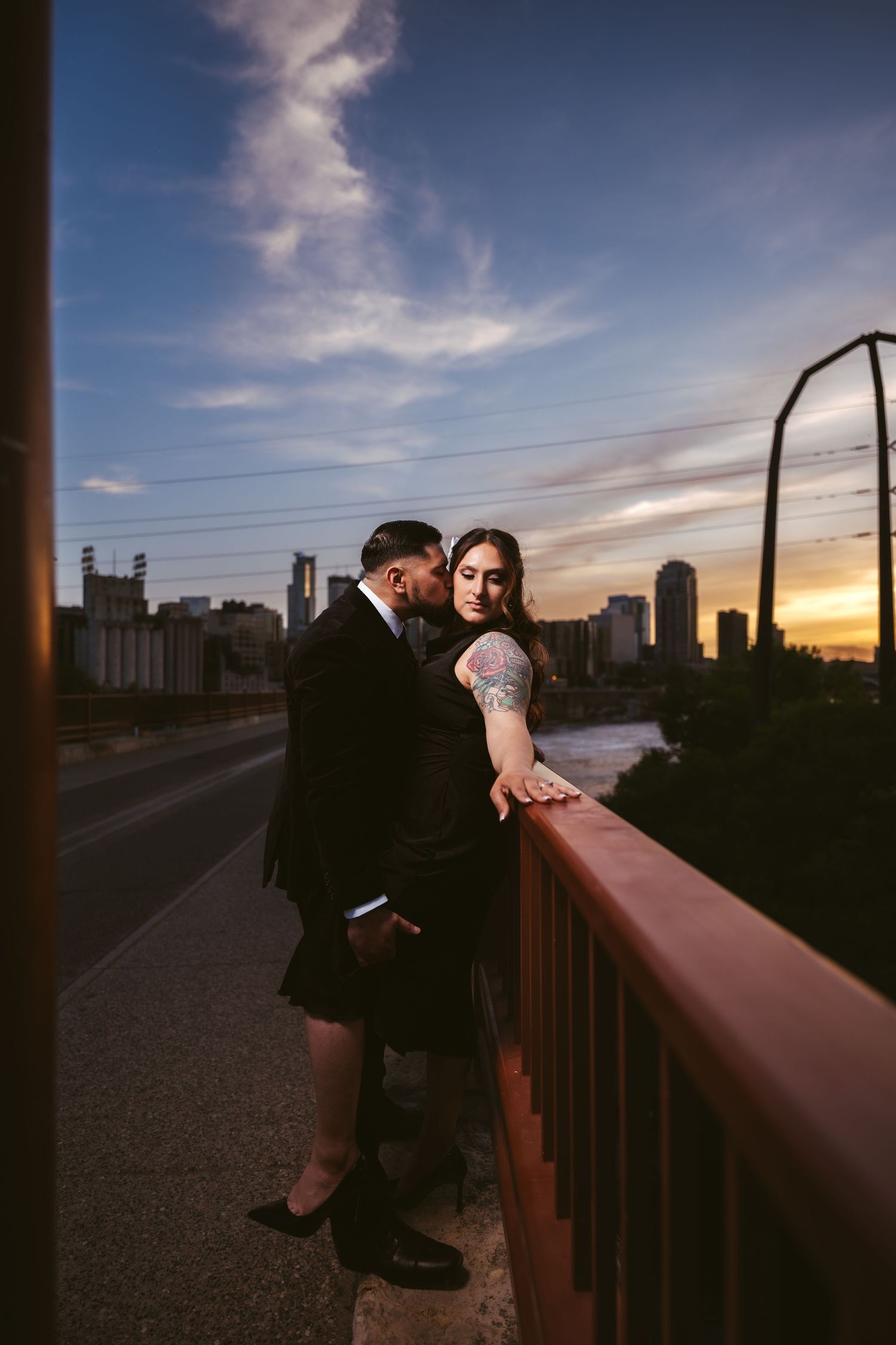 Couple in black formal wear sharing a tender moment on a bridge at sunset with city skyline views.