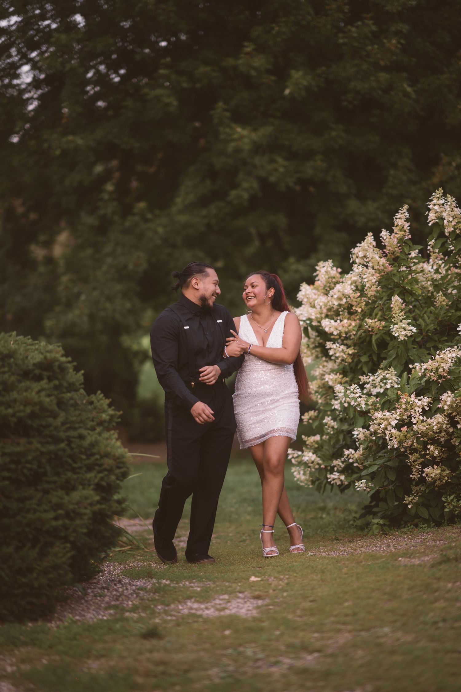 A couple poses playfully near blooming white flowers in a garden setting.